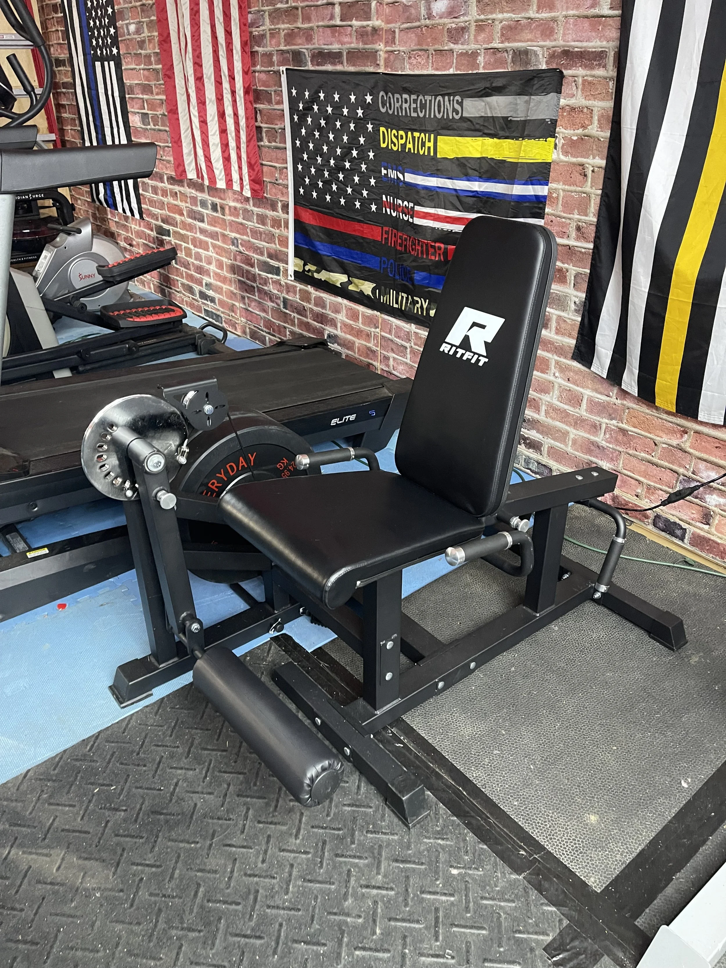 A seated calf raise machine in a gym with brick walls, fitness flags, and gym equipment around.