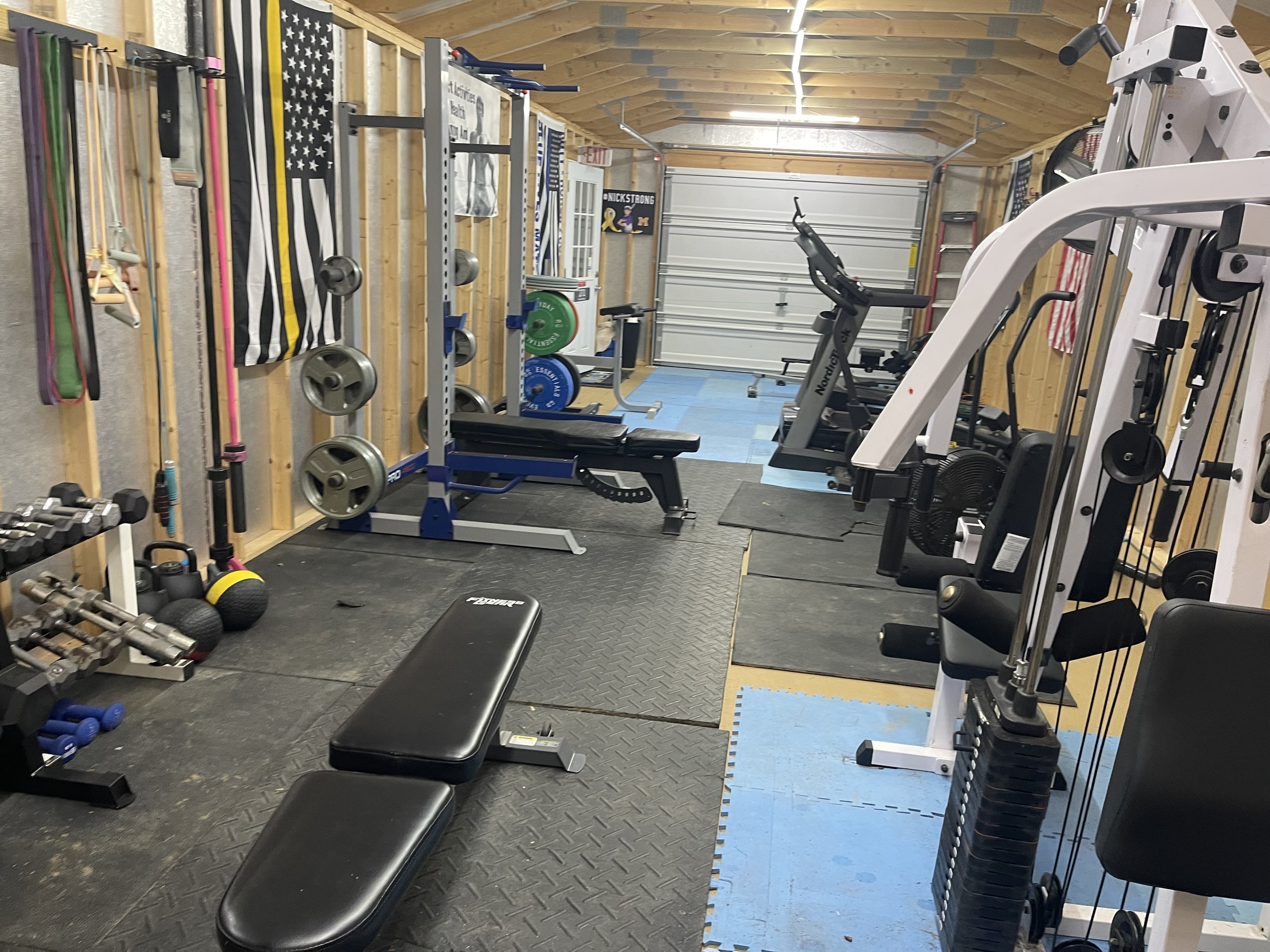 Home gym with weightlifting equipment, a weight bench, exercise bikes, and resistance bands on the wall, in a wooden-finished garage.