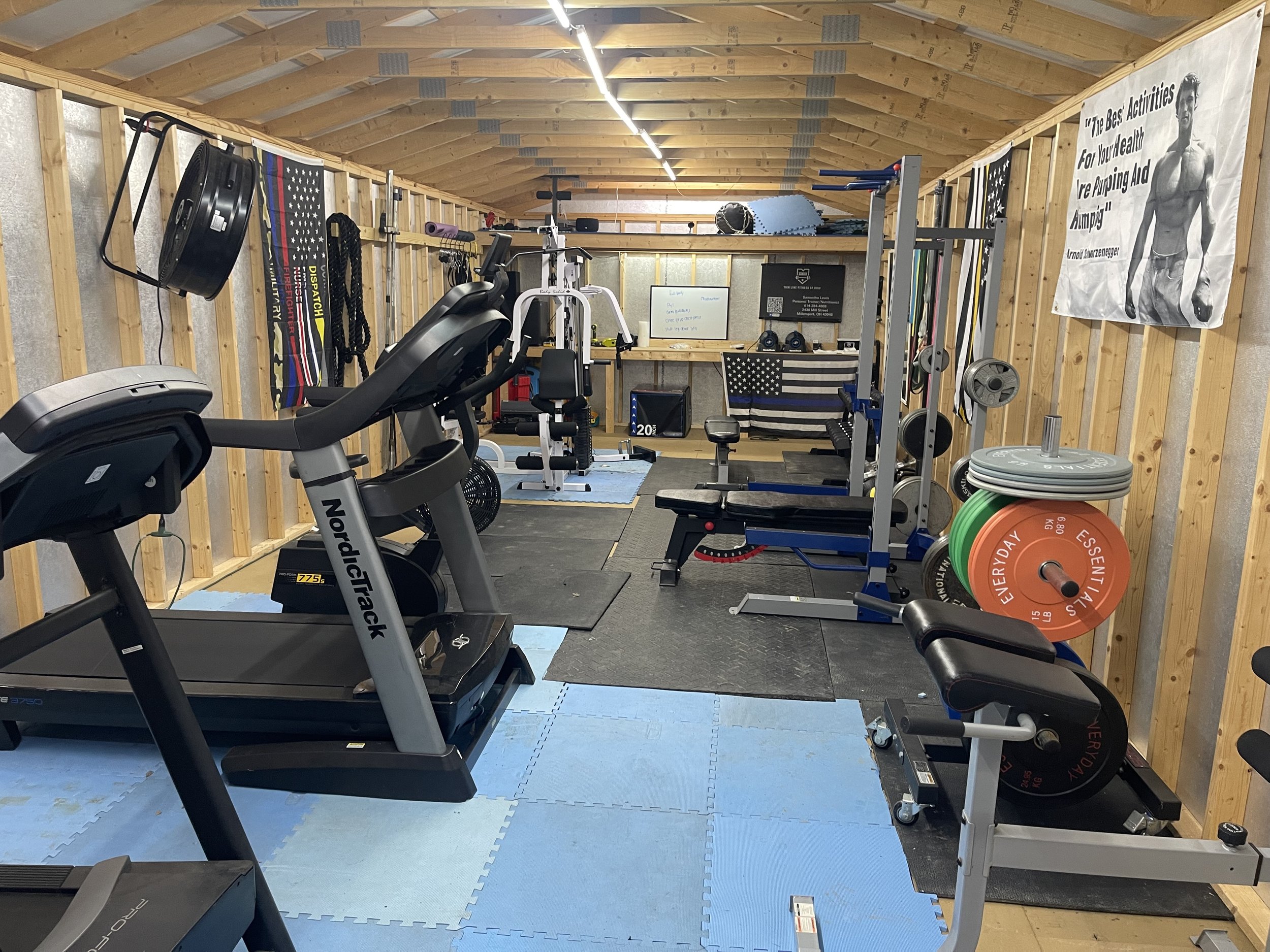 A home gym with various workout equipment including a treadmill, exercise bike, weight bench, weight plates, resistance bands, and a pull-up bar. The room has wooden walls and a sloped ceiling, with fitness posters and a small whiteboard on the back 