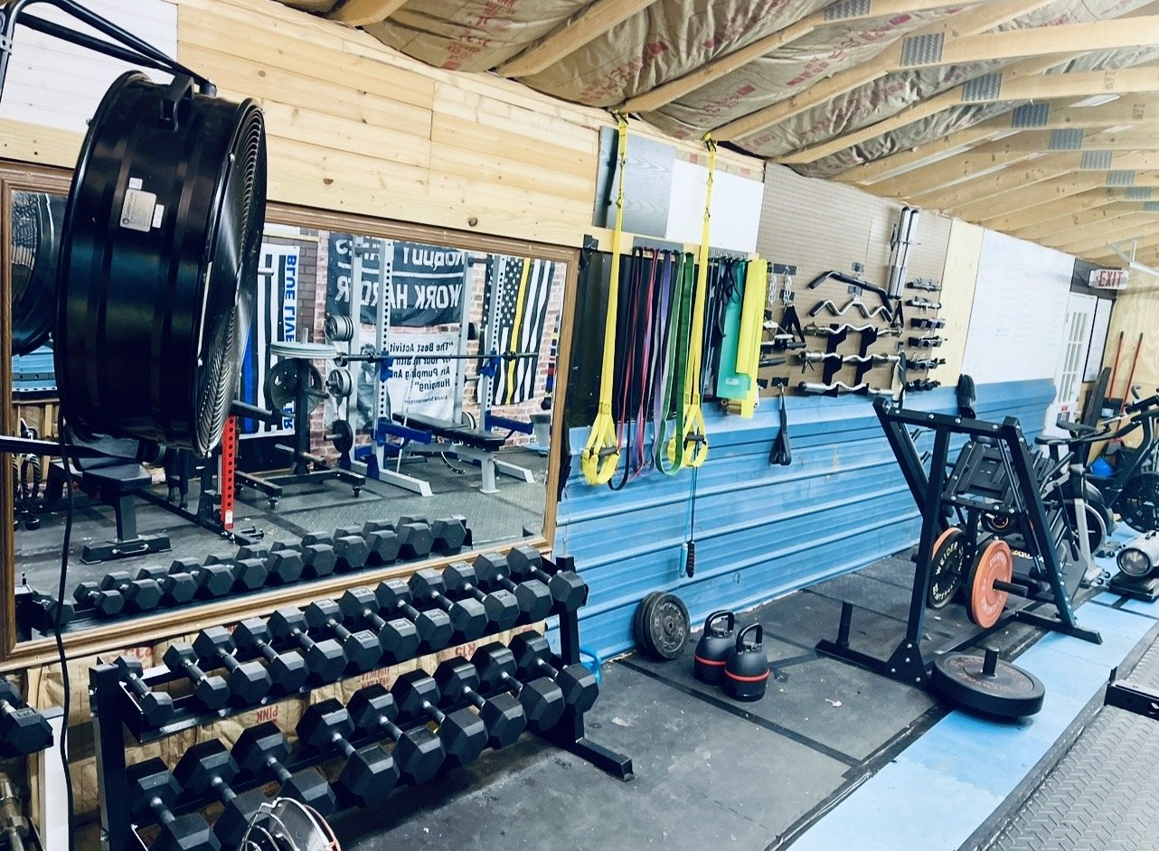 A gym with weightlifting equipment including dumbbell racks, barbells, kettlebells, resistance bands hanging on the wall, weight plates, a rowing machine, and a fan. The walls are made of wood, with a large mirror reflecting part of the gym, and the 