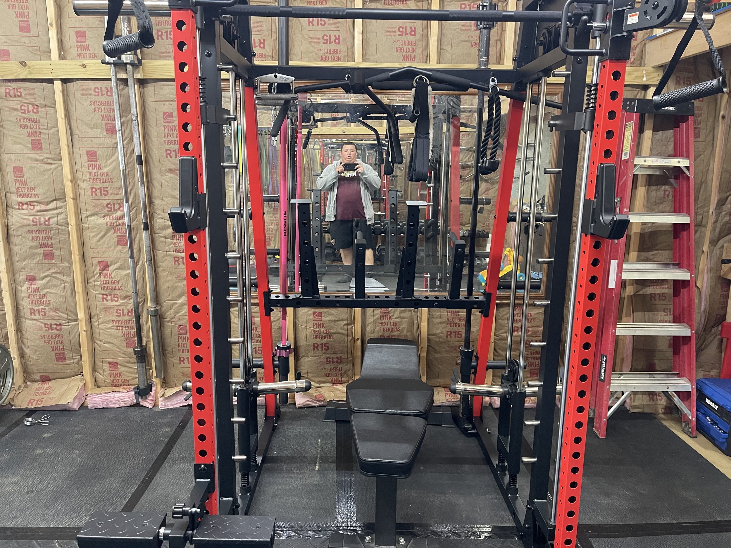 A home gym setup with a black and red power rack, a black adjustable weight bench, a ladder, and various gym equipment, with a large mirror on the wall and unfinished wood framing with insulation in the background.