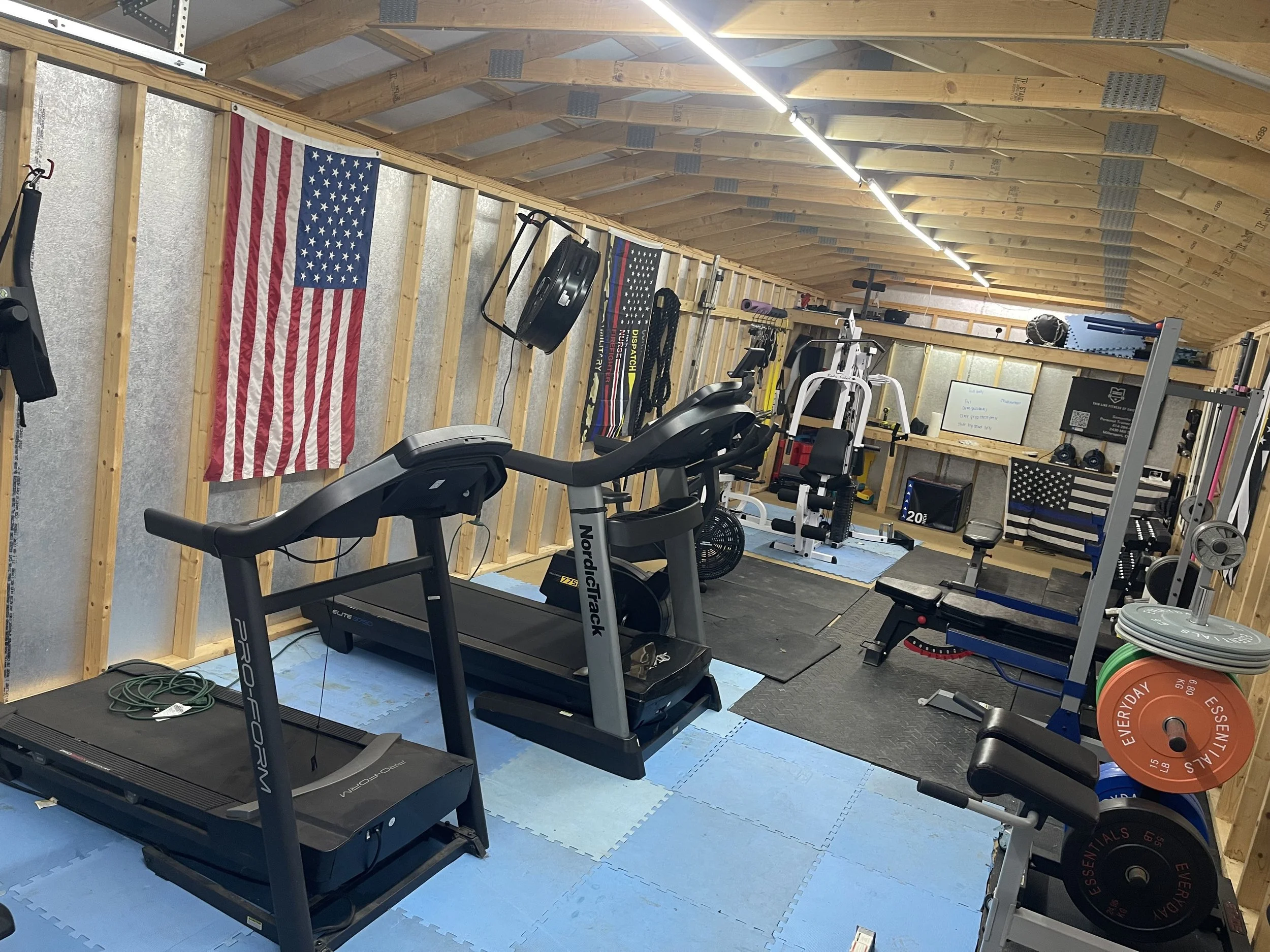 Home gym with American flags, treadmill, weightlifting equipment, and workout accessories in a wooden-finished room.