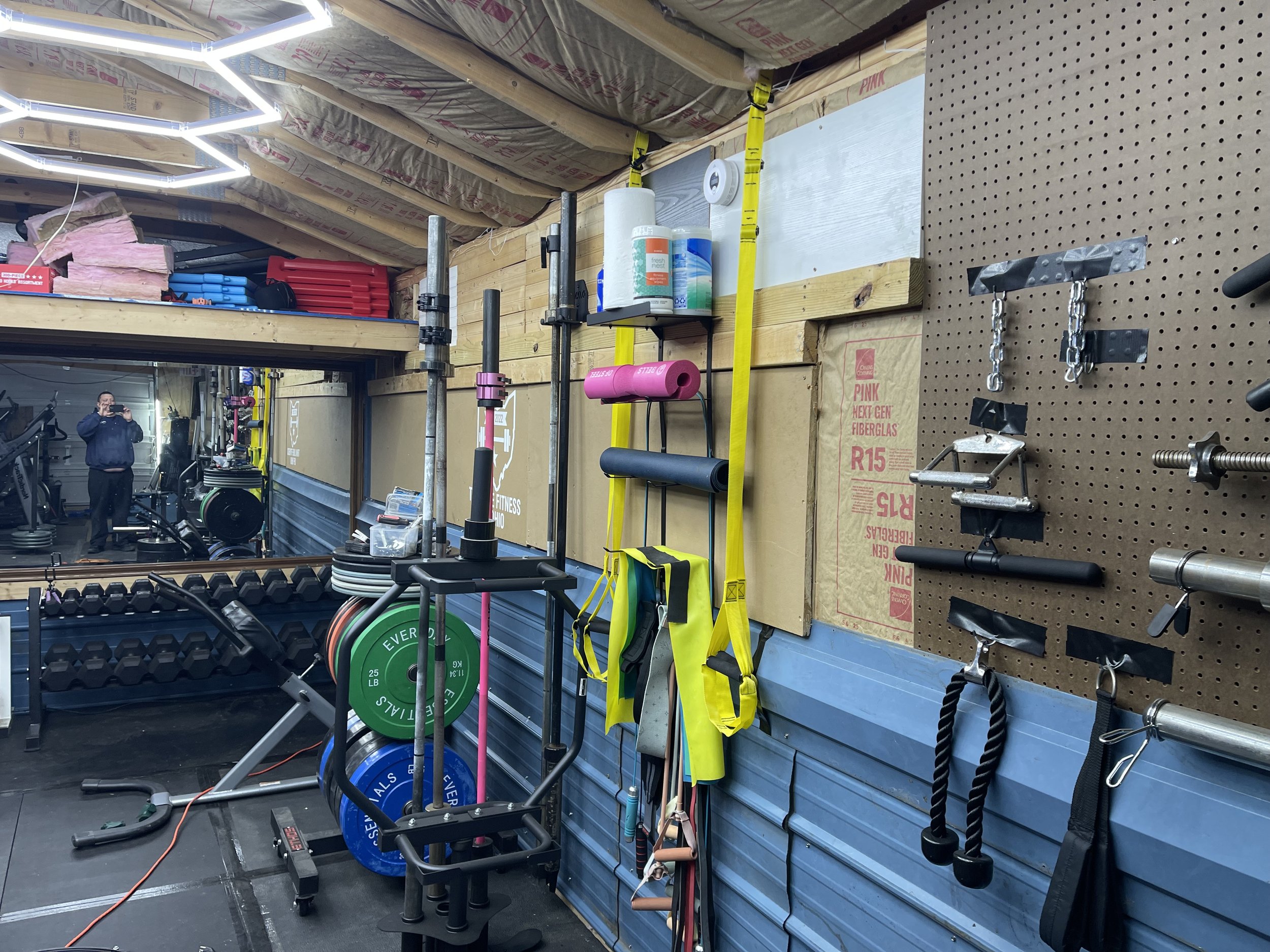 A gym with various fitness equipment and tools, including weight plates, barbells, resistance bands, and a pegboard with attachments, organized on the wall.