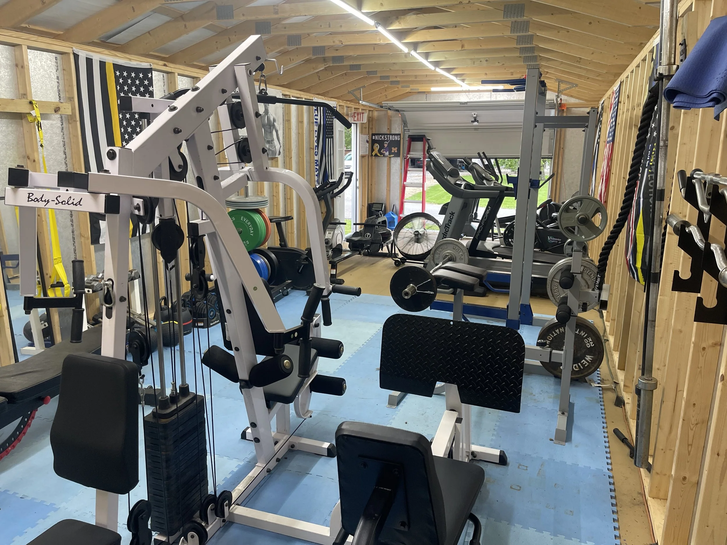 Home gym with various equipment including a white multi-function machine, a stationary bike, a treadmill, a weight rack, and free weights on the floor. The walls are covered with wooden paneling, and there are American flags and a motivational sign i