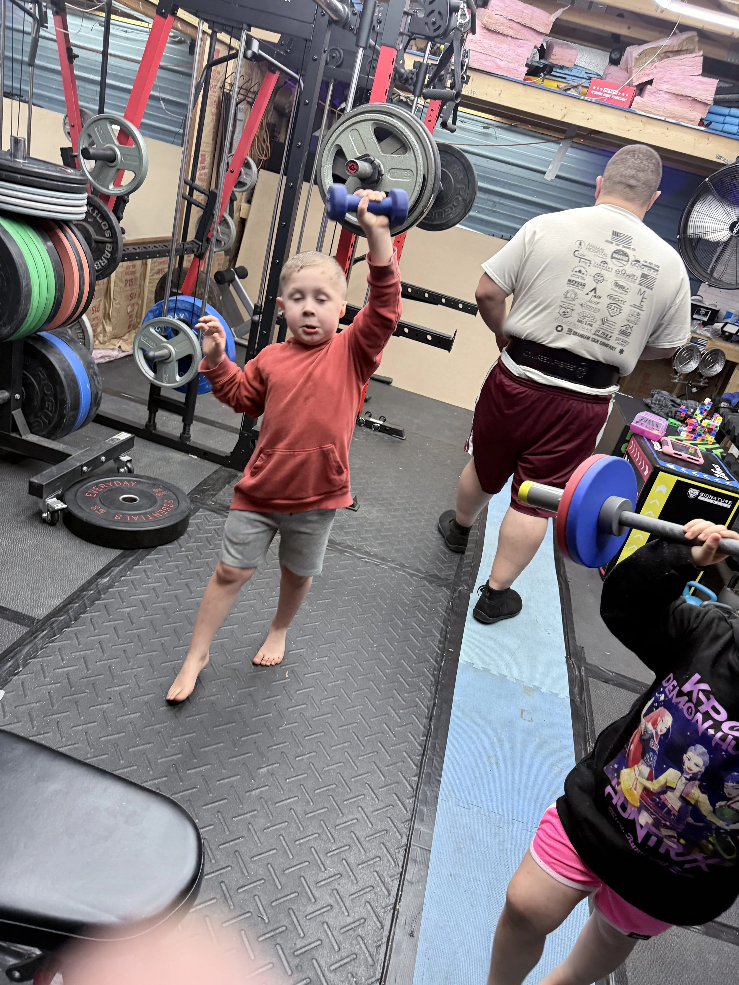 A young boy with blond hair, wearing a red hoodie and gray shorts, lifting a blue dumbbell in a gym. An adult man with a shaved head, wearing a light gray t-shirt and maroon shorts, is seen in the background, facing away from the camera. The gym has 