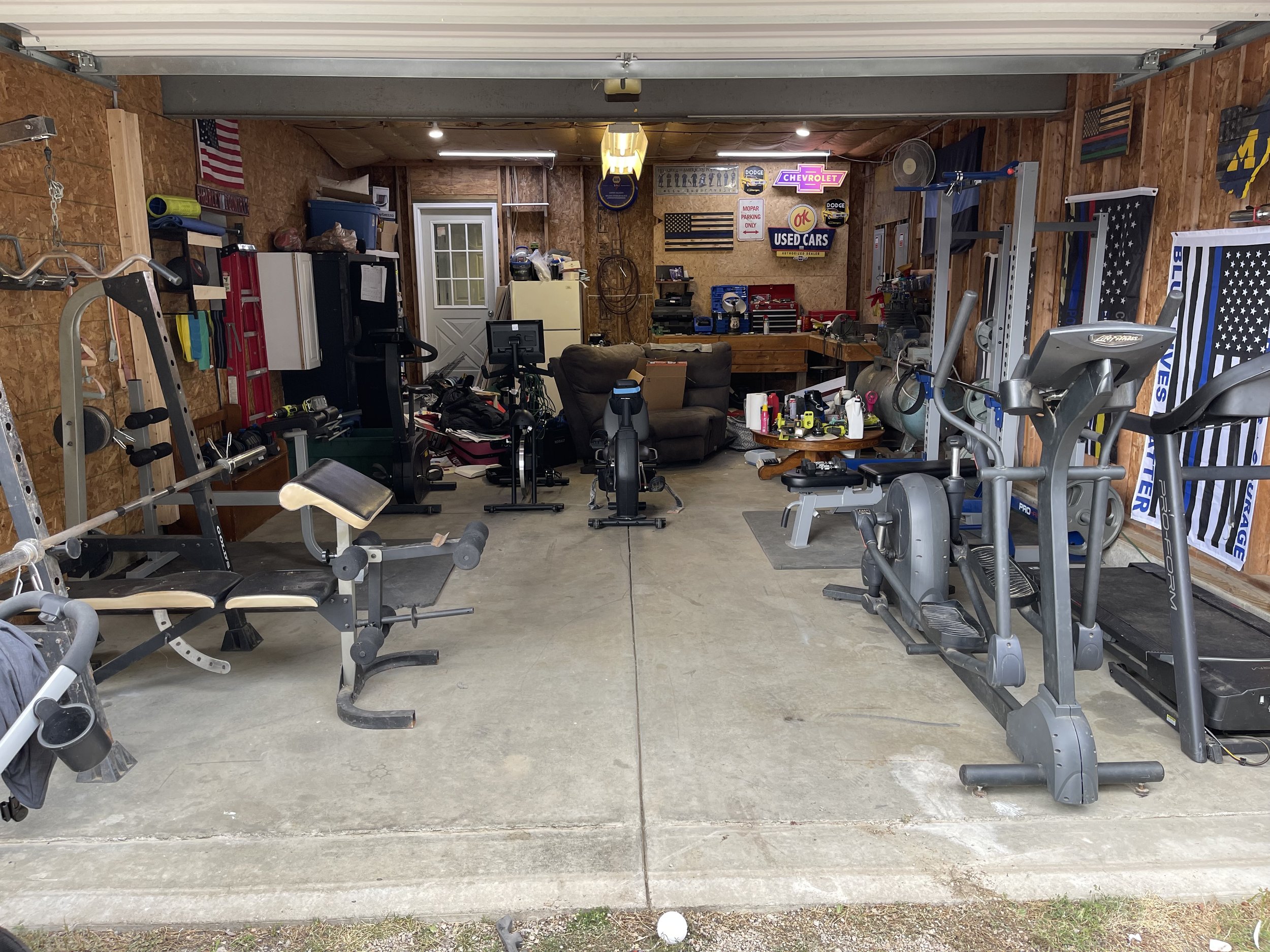 Garage with workout equipment, exercise bike, treadmill, weight machines, and flags on the wall, along with a seating area and various storage shelves.