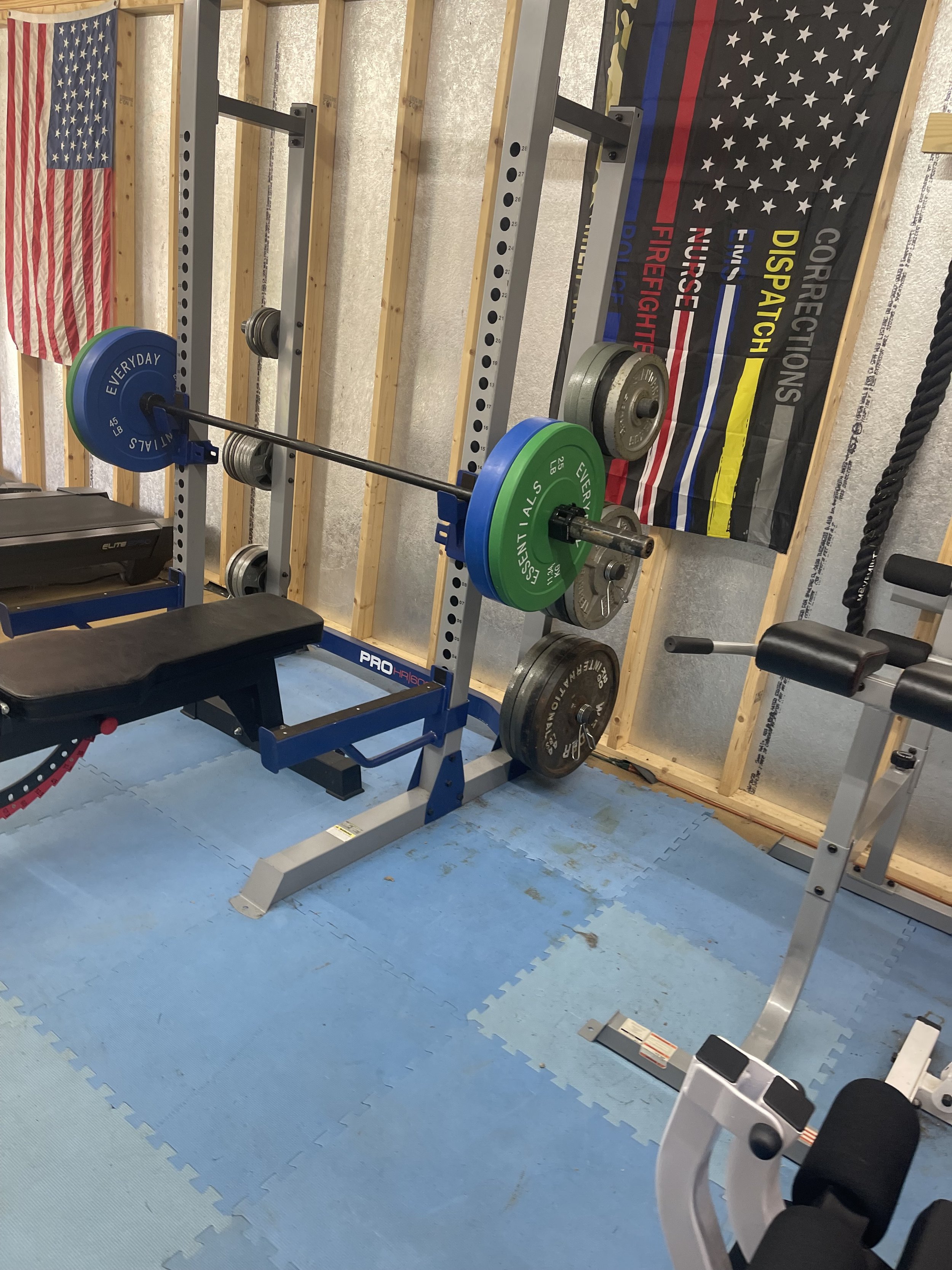 A home gym with a weightlifting bench, a squat rack with plates, American flags, and a gym banner on the wall.