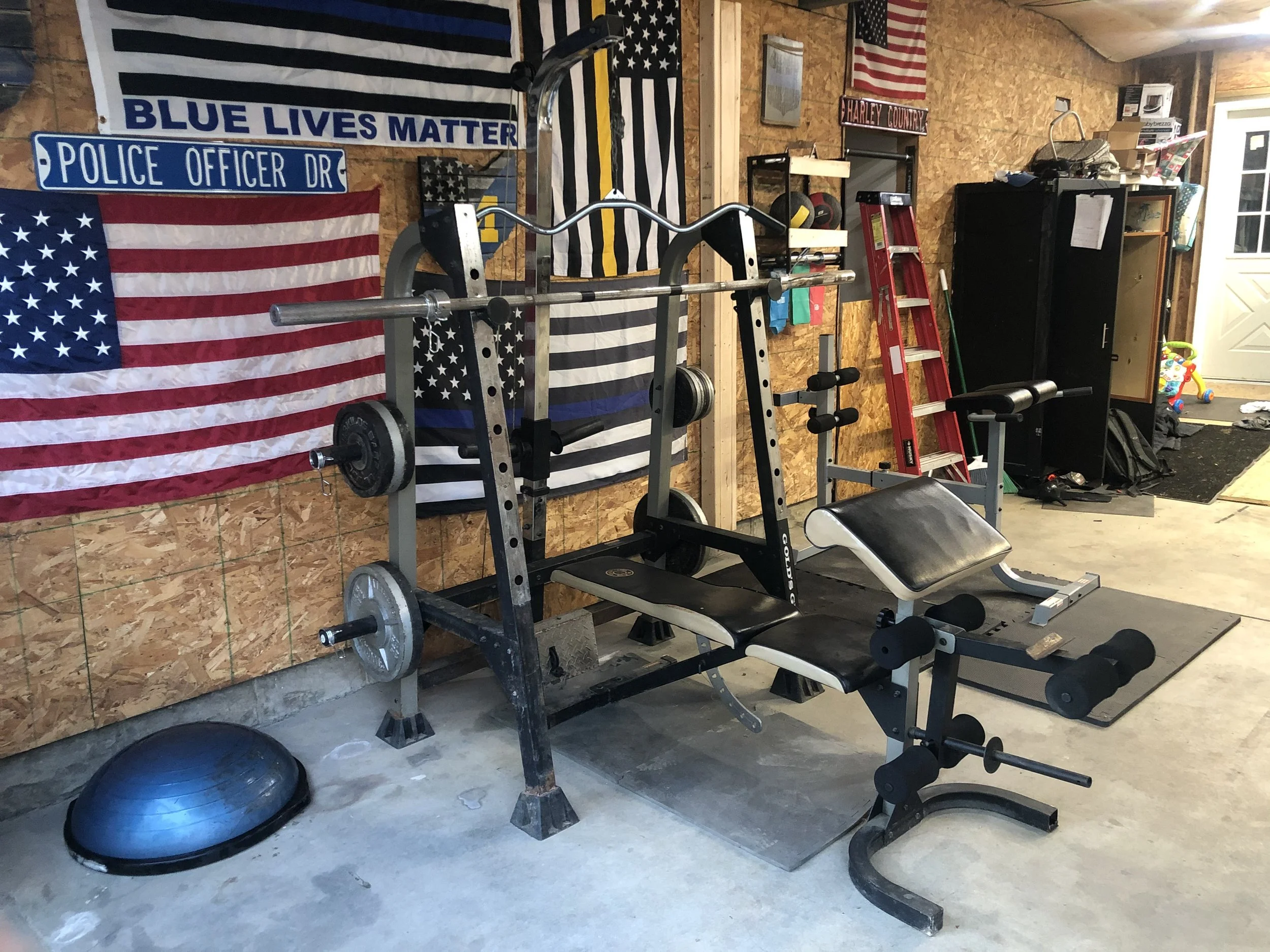 A garage workout area with exercise equipment, American flags, and banners, including a weight bench, barbell, and a stability ball on a concrete floor.