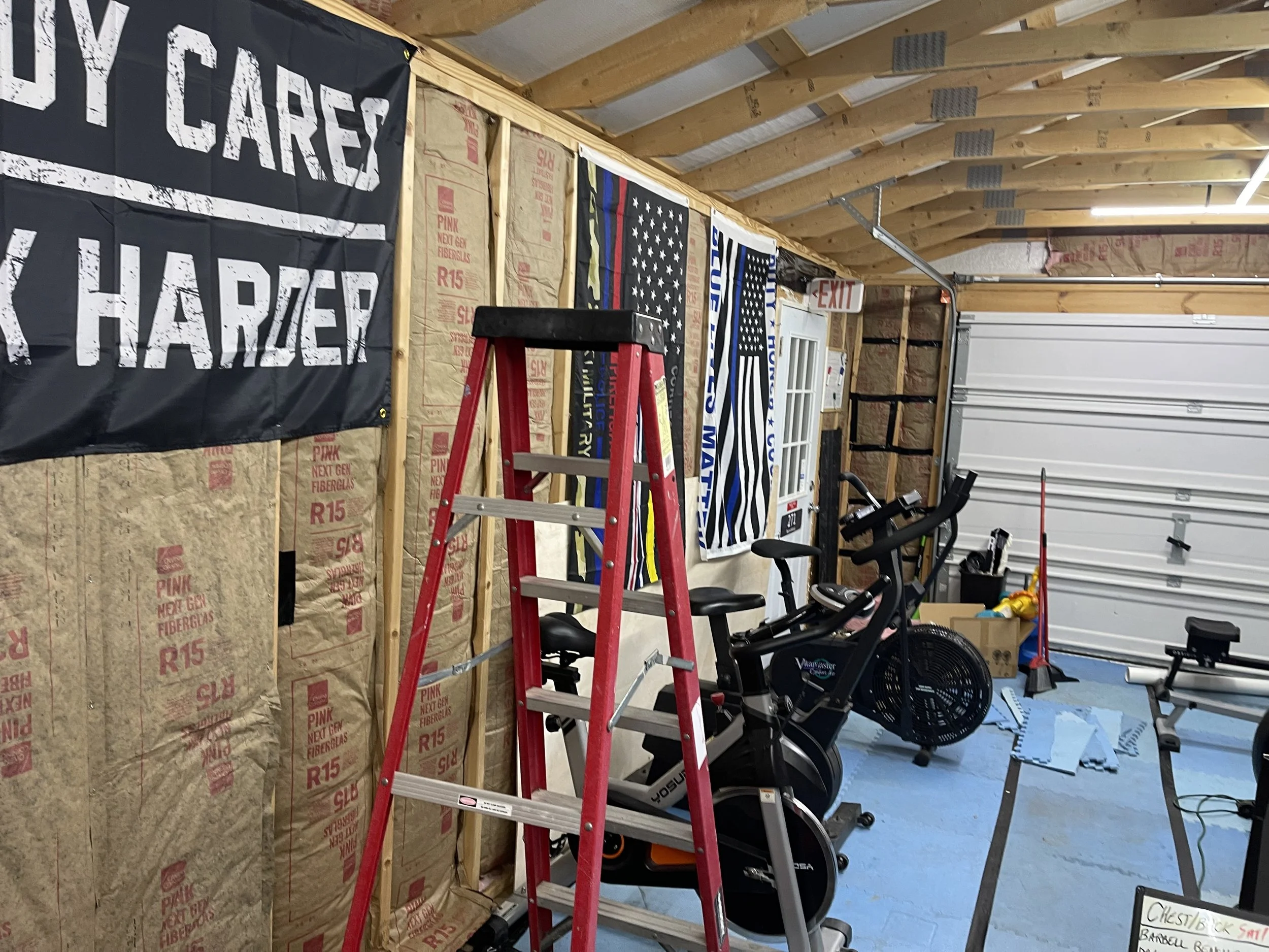 A garage with insulation and wood framing, containing exercise bikes, a ladder, cleaning tools, and patriotic banners hanging on the wall.