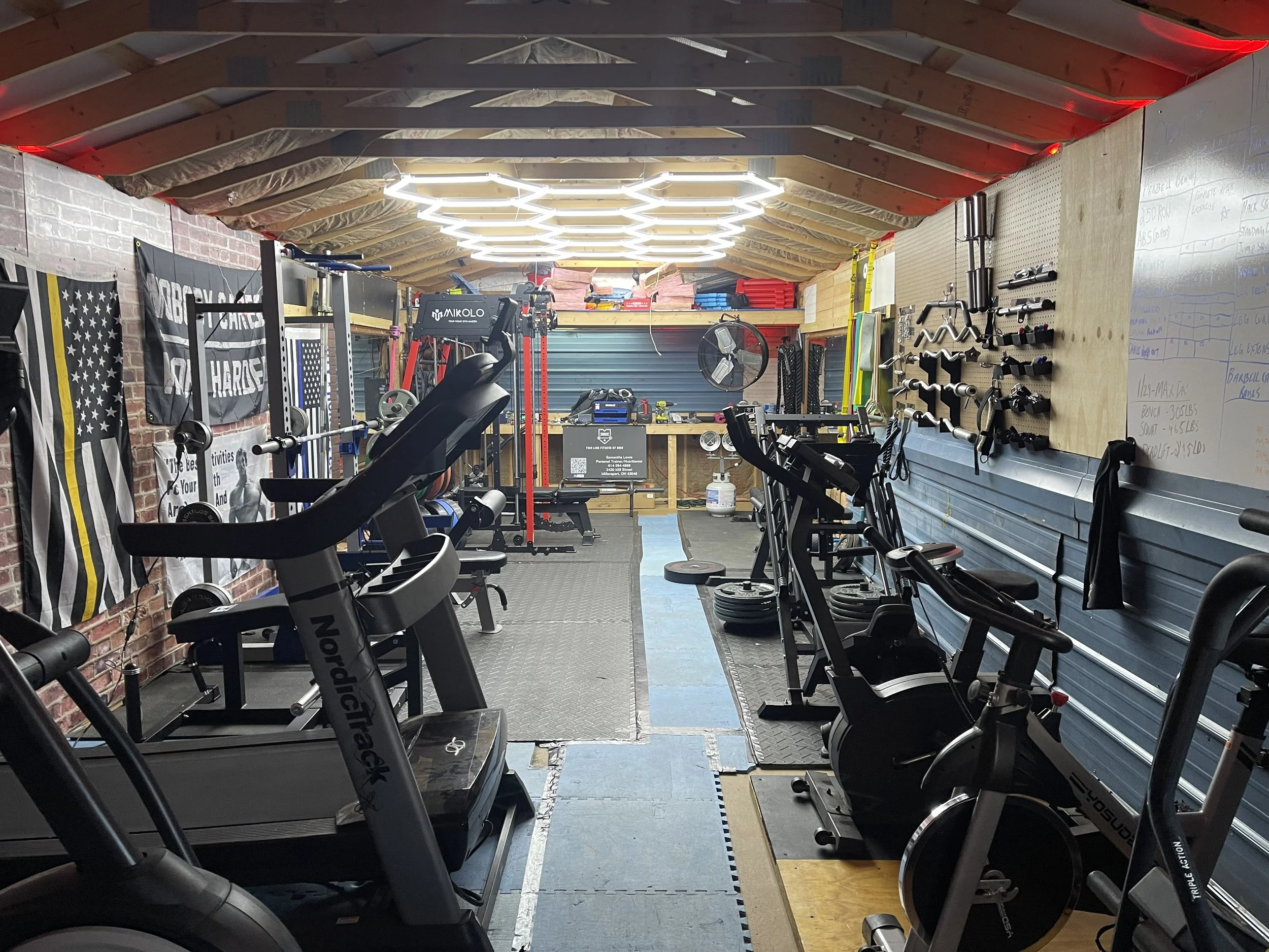 A home gym with various fitness equipment including treadmills, stationary bikes, and weights, with American flags hanging on the brick wall.
