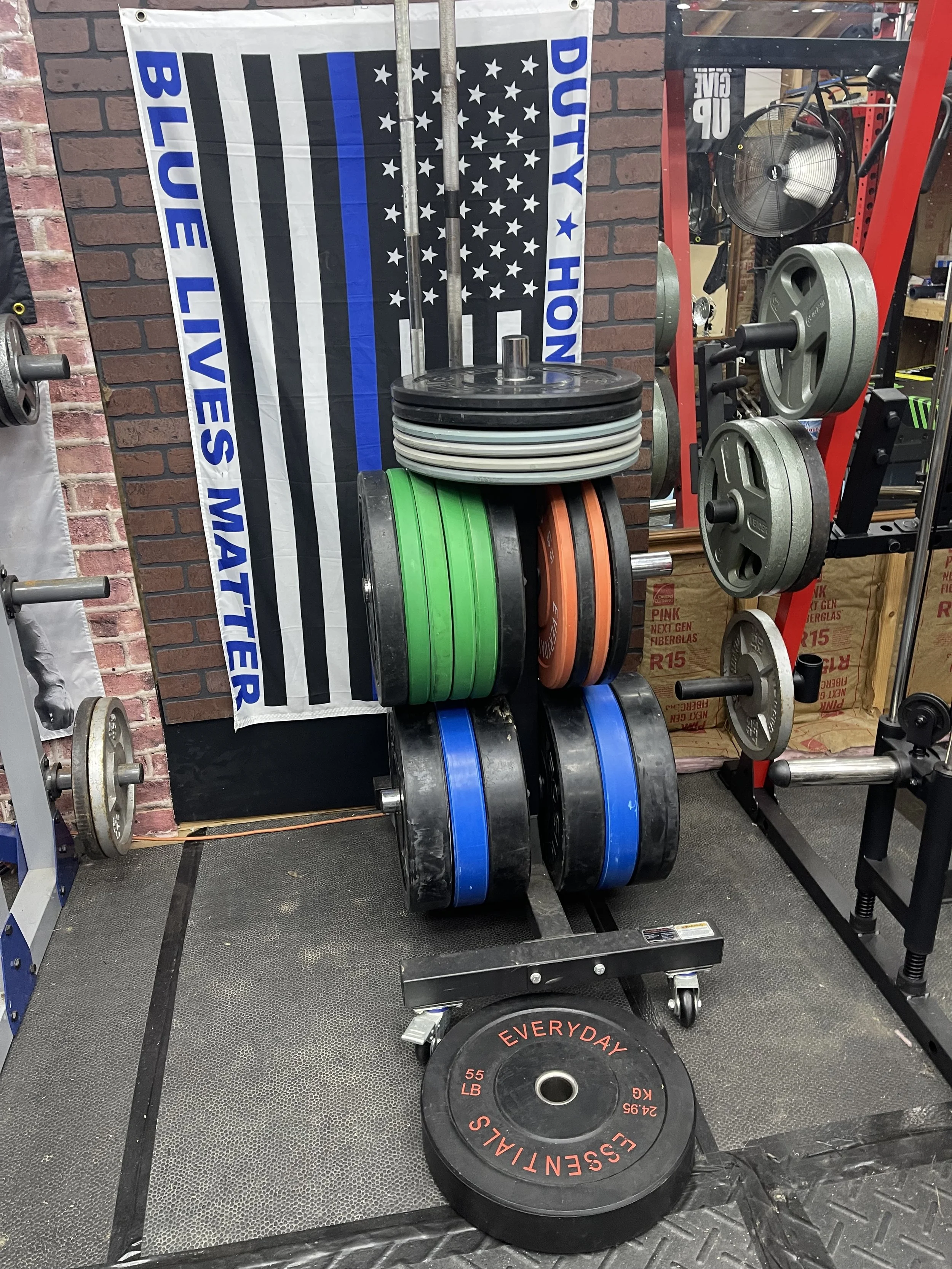 A weightlifting rack with various weight plates stacked on it, with a large black weight plate in the foreground labeled "EVERYDAY ESSENTIALS." A banner with a black and white American flag with a blue stripe and the words "DUTY HONOR BLUE LIVES MATT