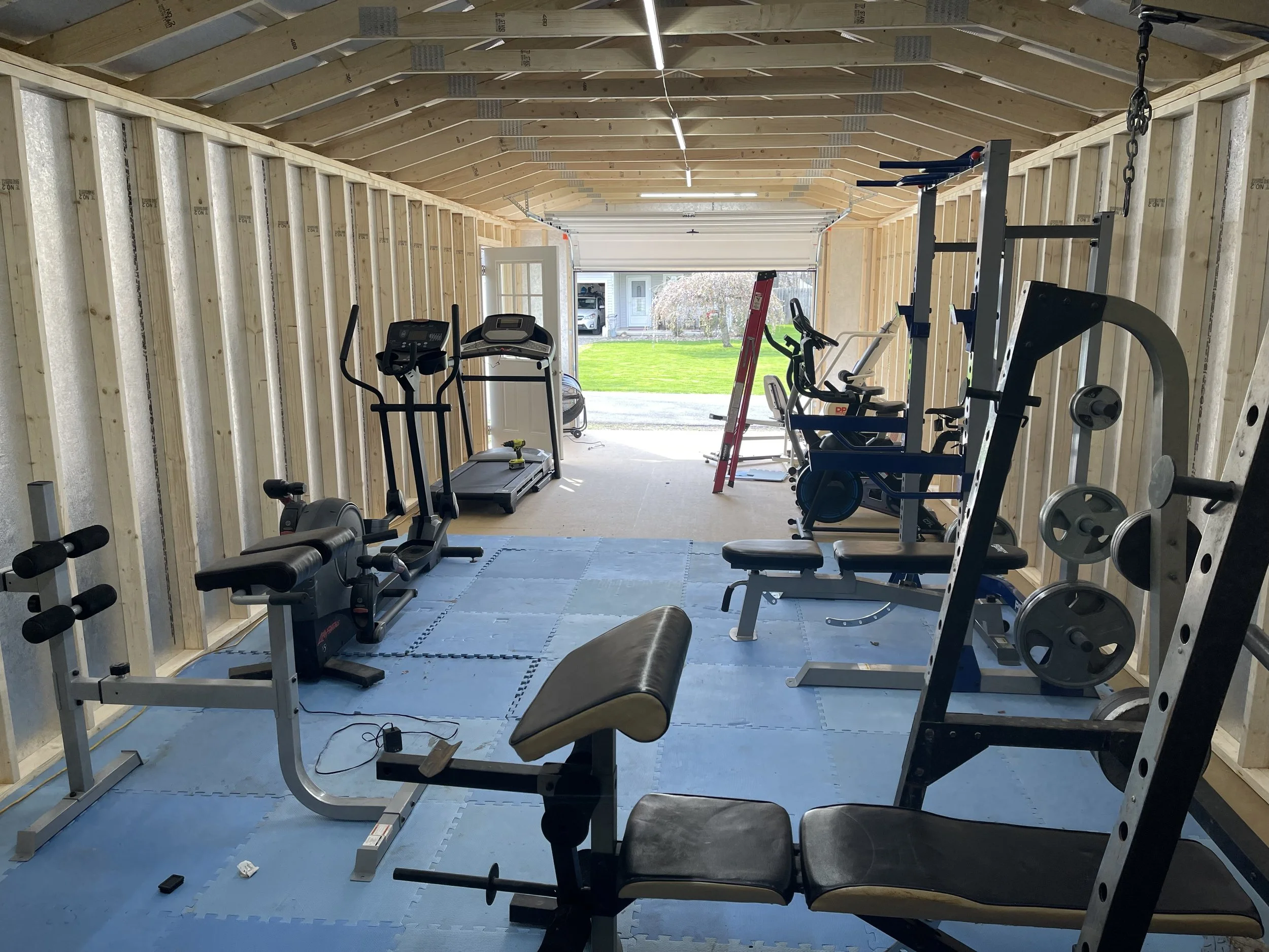 Inside a partially built home gym with wooden framing and blue interlocking foam mats on the floor. The gym contains various workout equipment, including a treadmill, elliptical machine, stationary bike, weight bench with barbell, and a squat rack wi