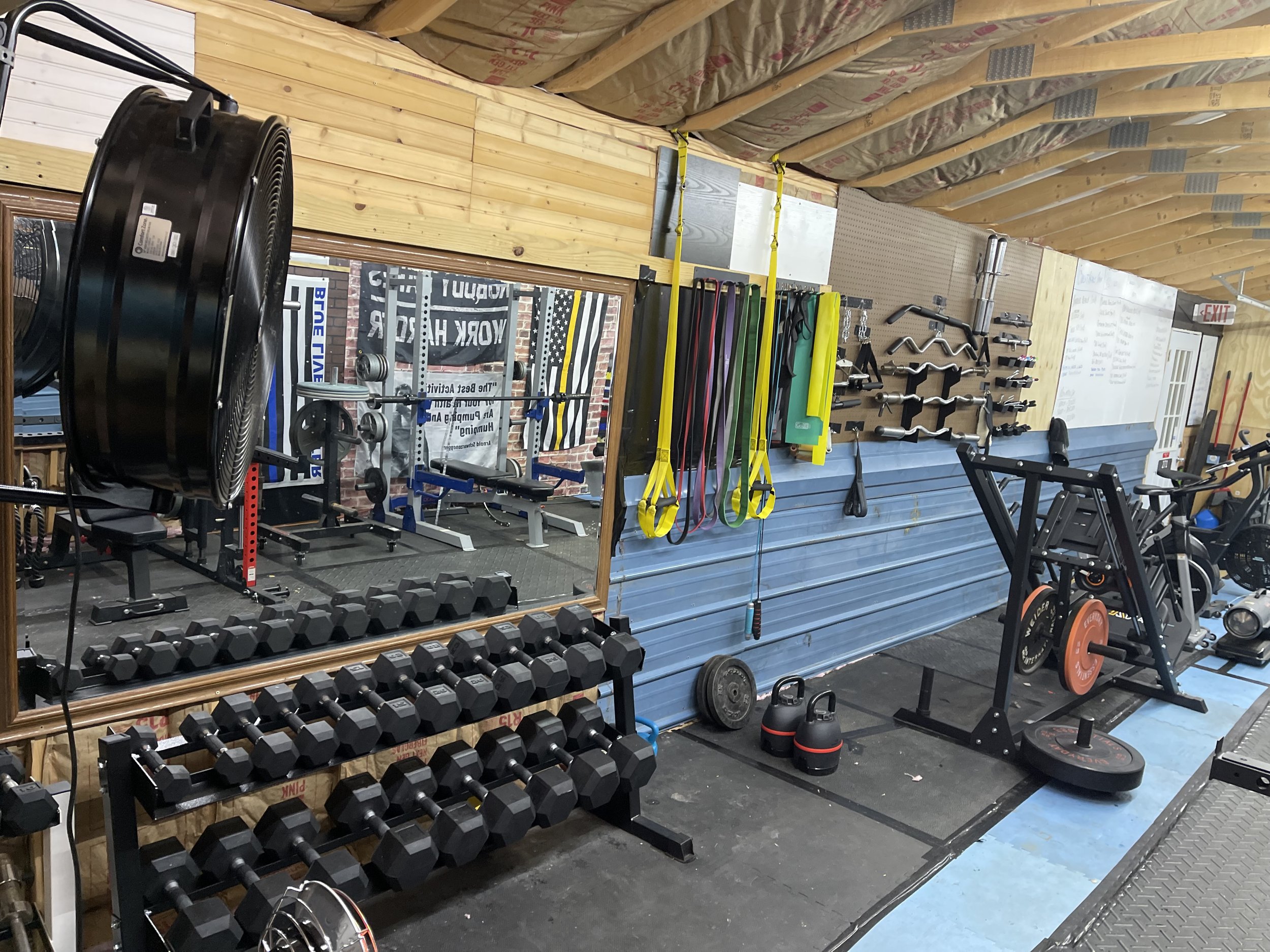 Inside a gym with kettlebells, dumbbells, weight plates, resistance bands, pull-up bars, and various fitness equipment against wooden walls, with a mirror reflecting more workout stations.