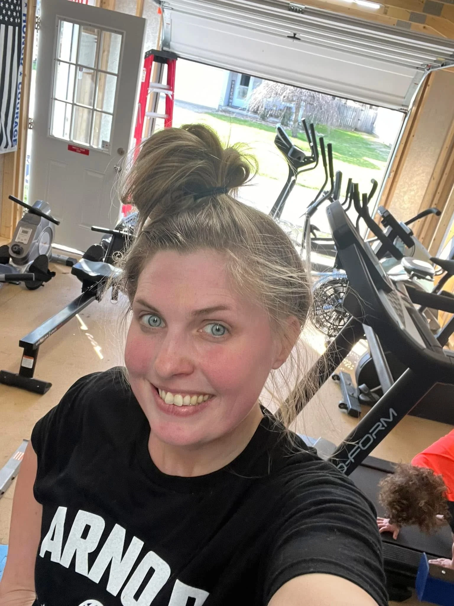 A woman taking a selfies inside a home gym with exercise equipment, including rowing machines, in the background, and a child's hand and head visible behind her.