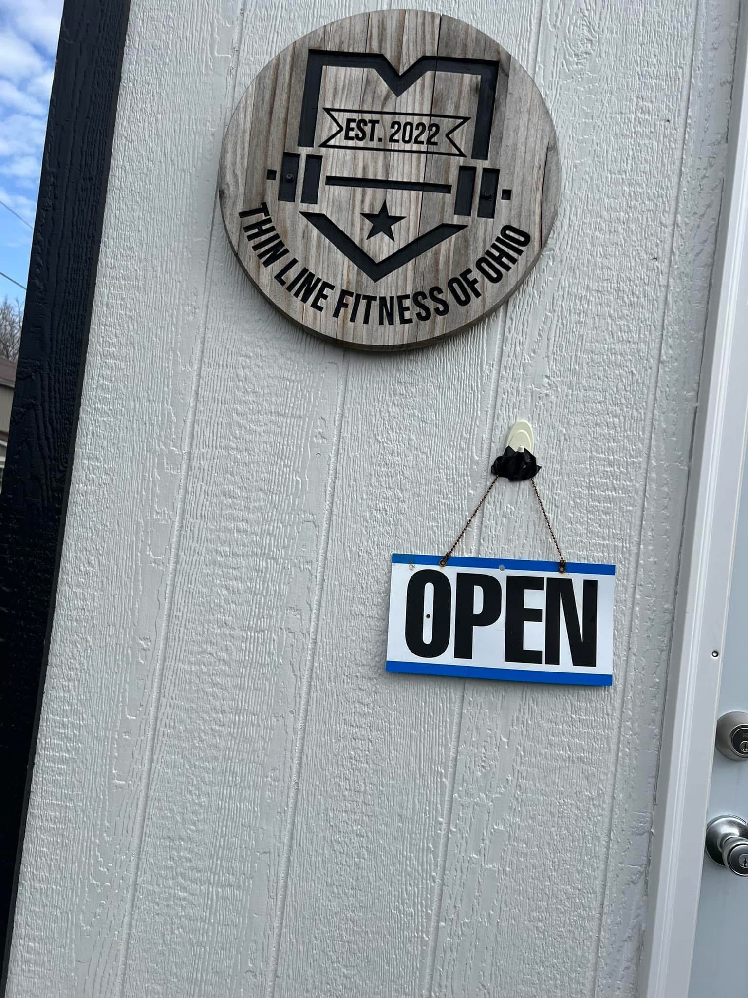 Sign on a door with a round wooden plaque reading "The Lin Fitness of Ohio," established in 2022, and an attached "Open" sign.