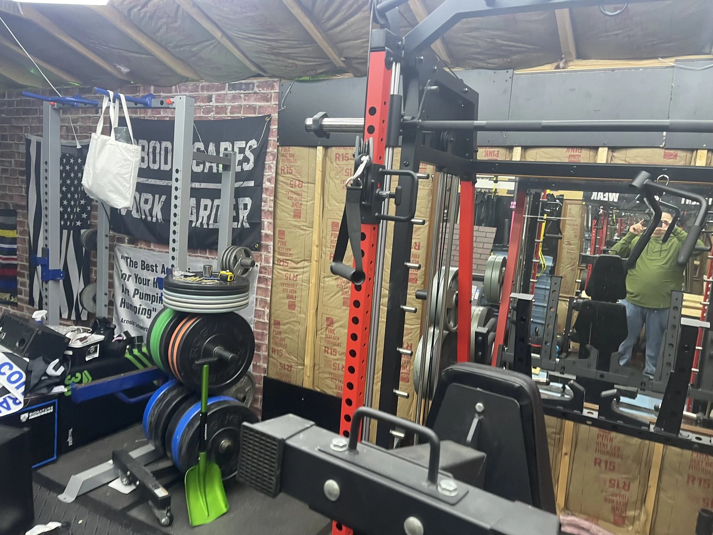 Gym workout area with weightlifting equipment, barbells, weight plates, a mirror, a person wearing a green hoodie, and various workout accessories.
