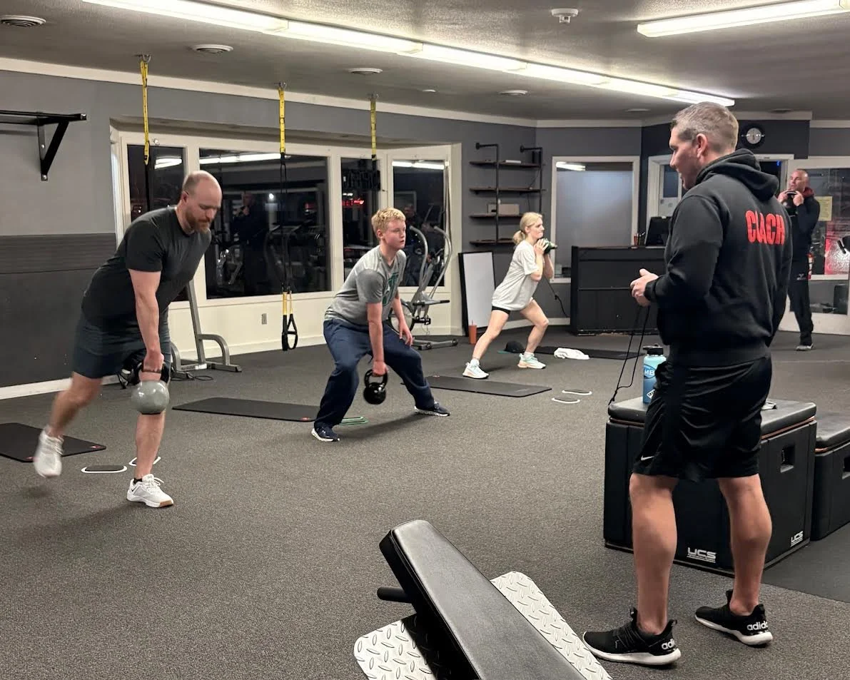 Four people exercising with kettlebells and dumbbells in a gym, a trainer supervising them, and others in the background.