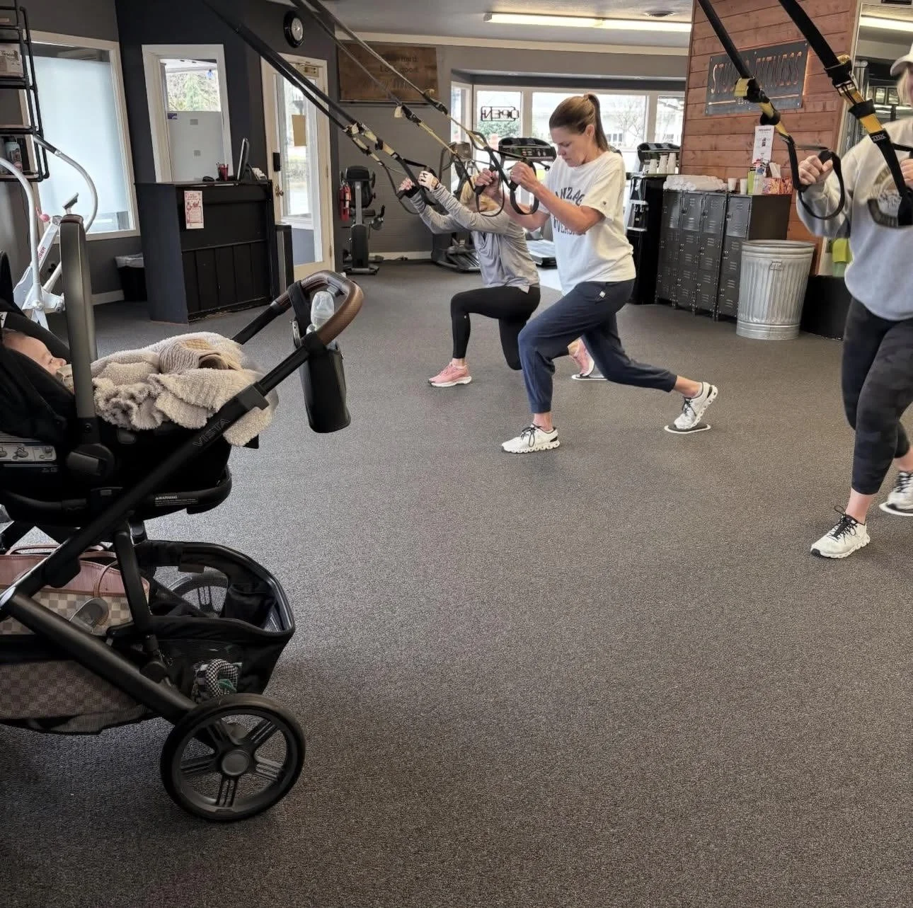 Four women exercising with TRX suspension straps in a gym, one woman is using a stroller with a sleeping baby, and others are doing squats.