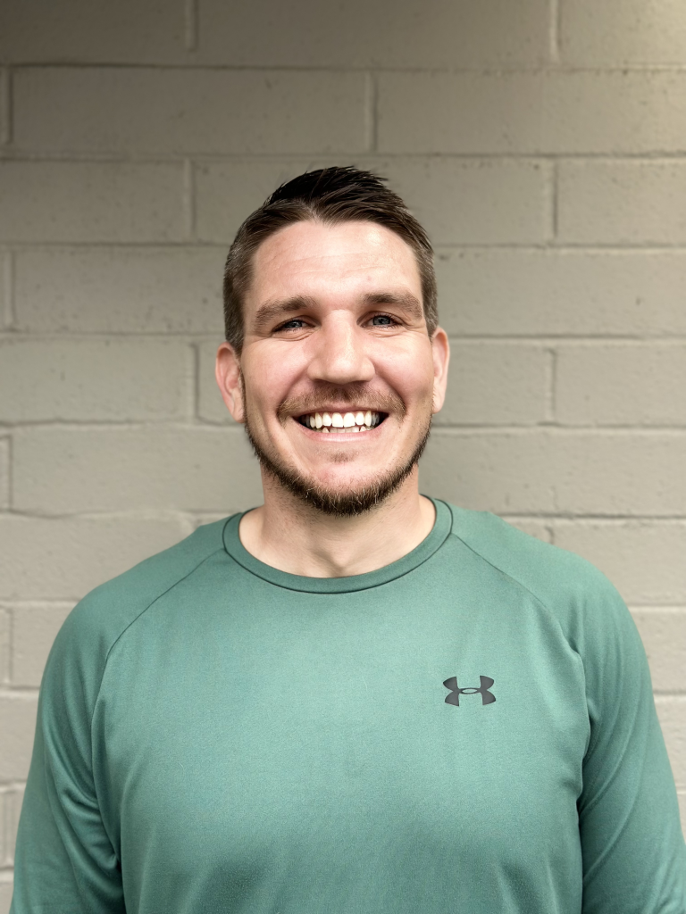 Smiling man with short brown hair and a beard wearing a green long-sleeve athletic shirt standing in front of a beige brick wall.