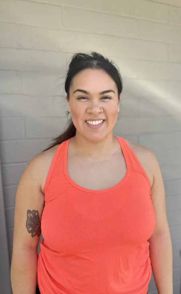 A smiling woman with dark hair in a ponytail, wearing a bright orange athletic tank top, standing outdoors against a light gray brick wall, showing a tattoo on her left arm.