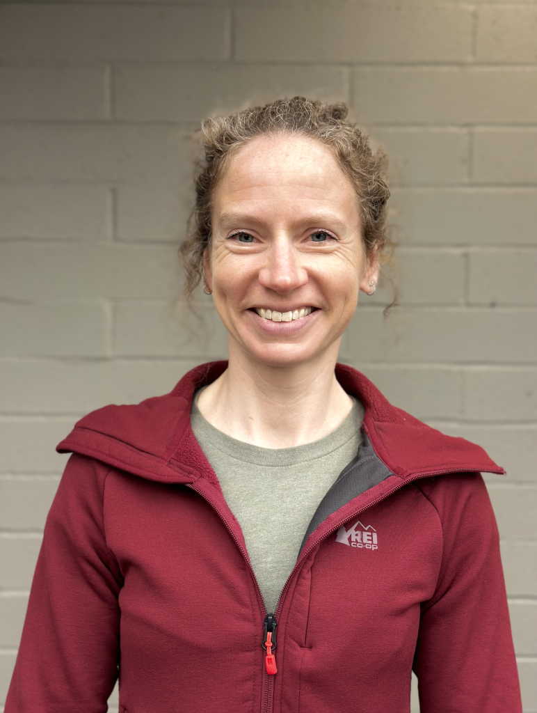 Smiling woman with curly hair wearing a maroon jacket and gray shirt in front of a beige brick wall.