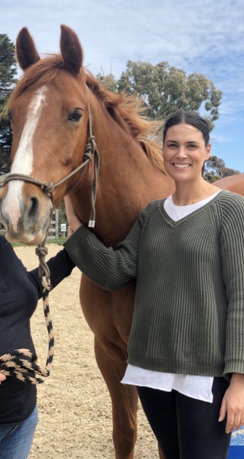 A woman smiling and standing next to a brown horse with a white stripe on its face, outdoors on a sunny day with trees and blue sky in the background.