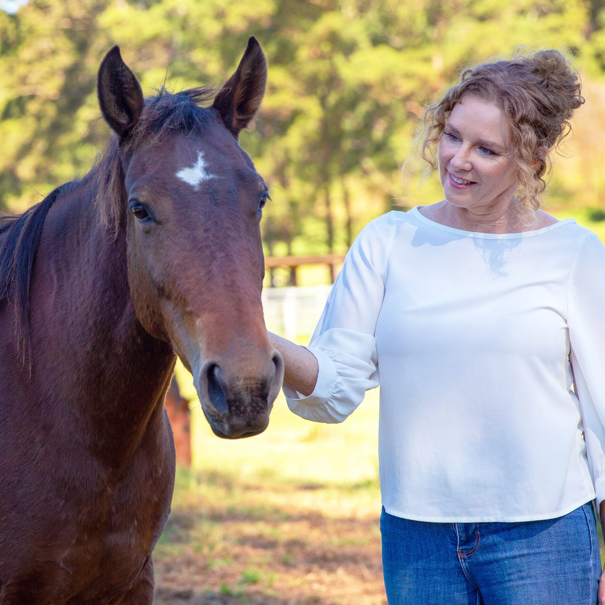 A woman with curly blonde hair wearing a white top and jeans standing outdoors near a brown horse with a white star on its forehead, surrounded by green trees and sunlight.