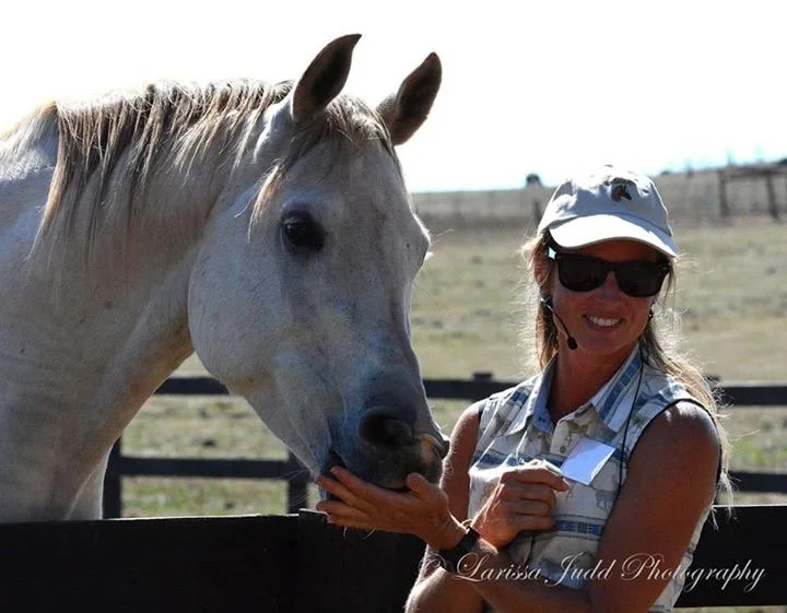 A woman wearing sunglasses, a white cap, and a sleeveless vest petting a white horse near a wooden fence on a sunny day.