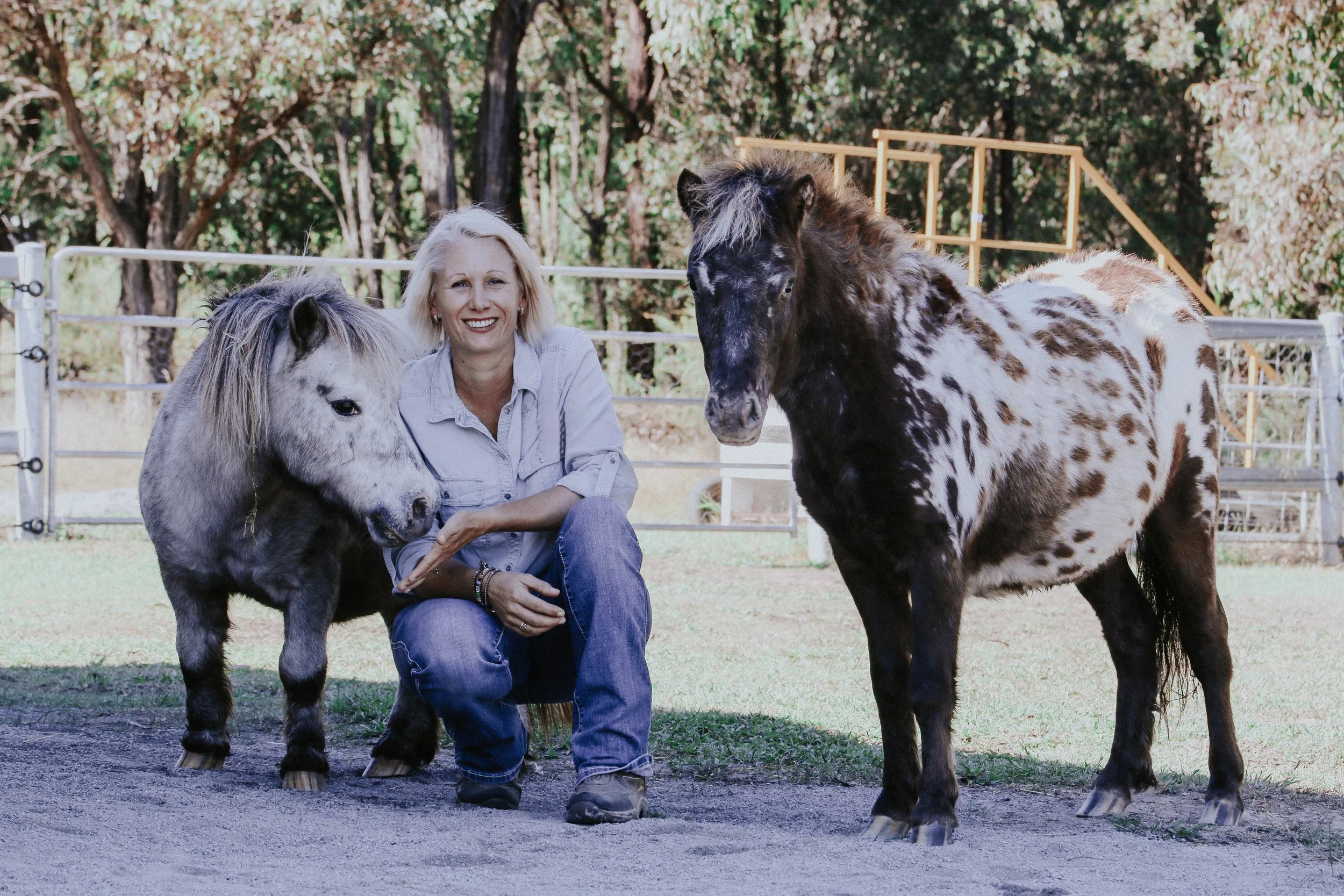 A woman smiles as she kneels next to two young ponies in an outdoor paddock with trees and a metal fence in the background.