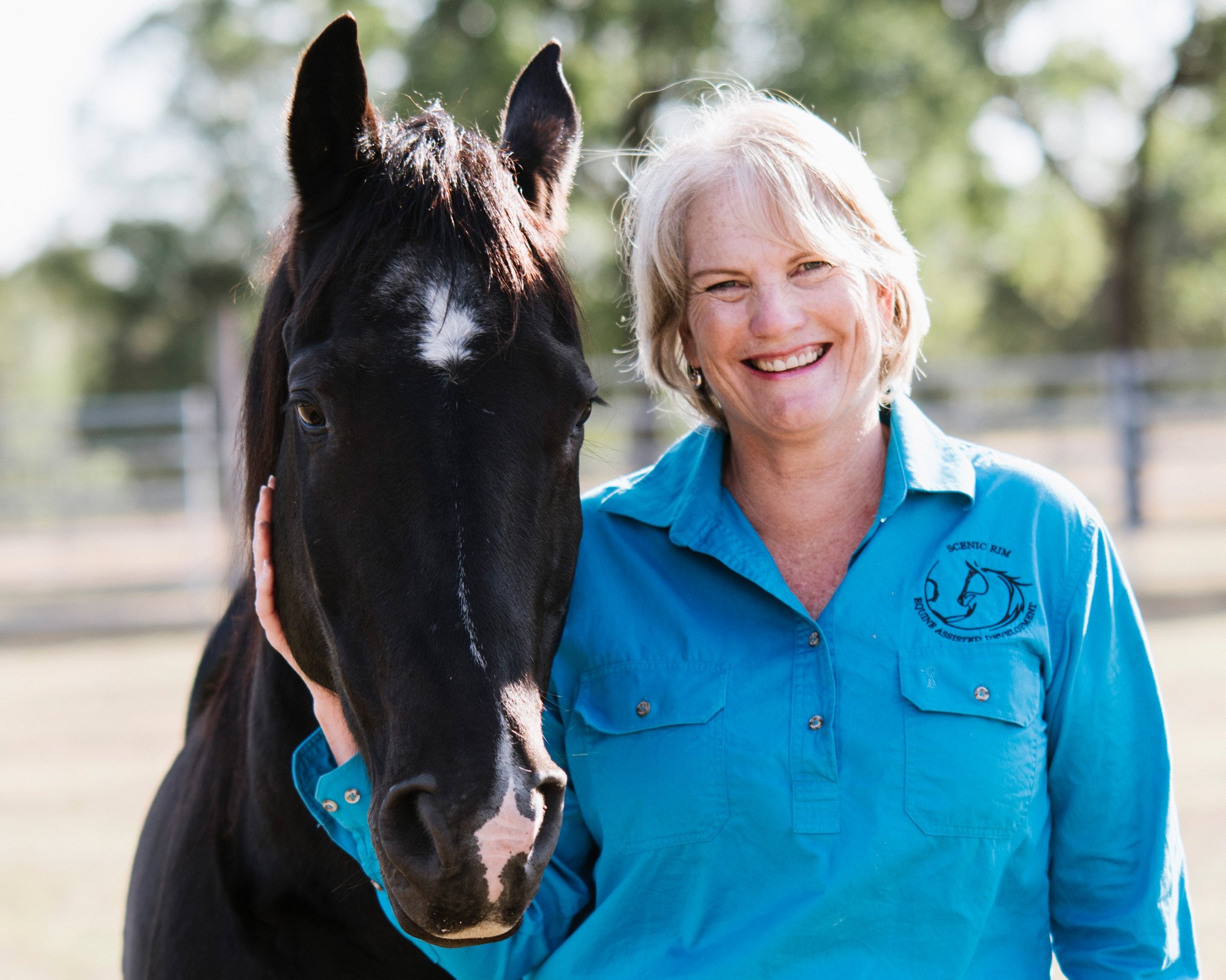 A woman with short blonde hair and a blue shirt smiling next to a black horse outdoors.