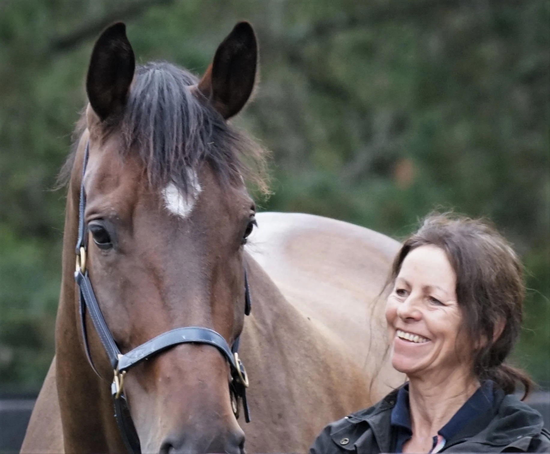 A woman smiling stands next to a brown horse with a white star on its forehead, both outdoors with a blurred green background.
