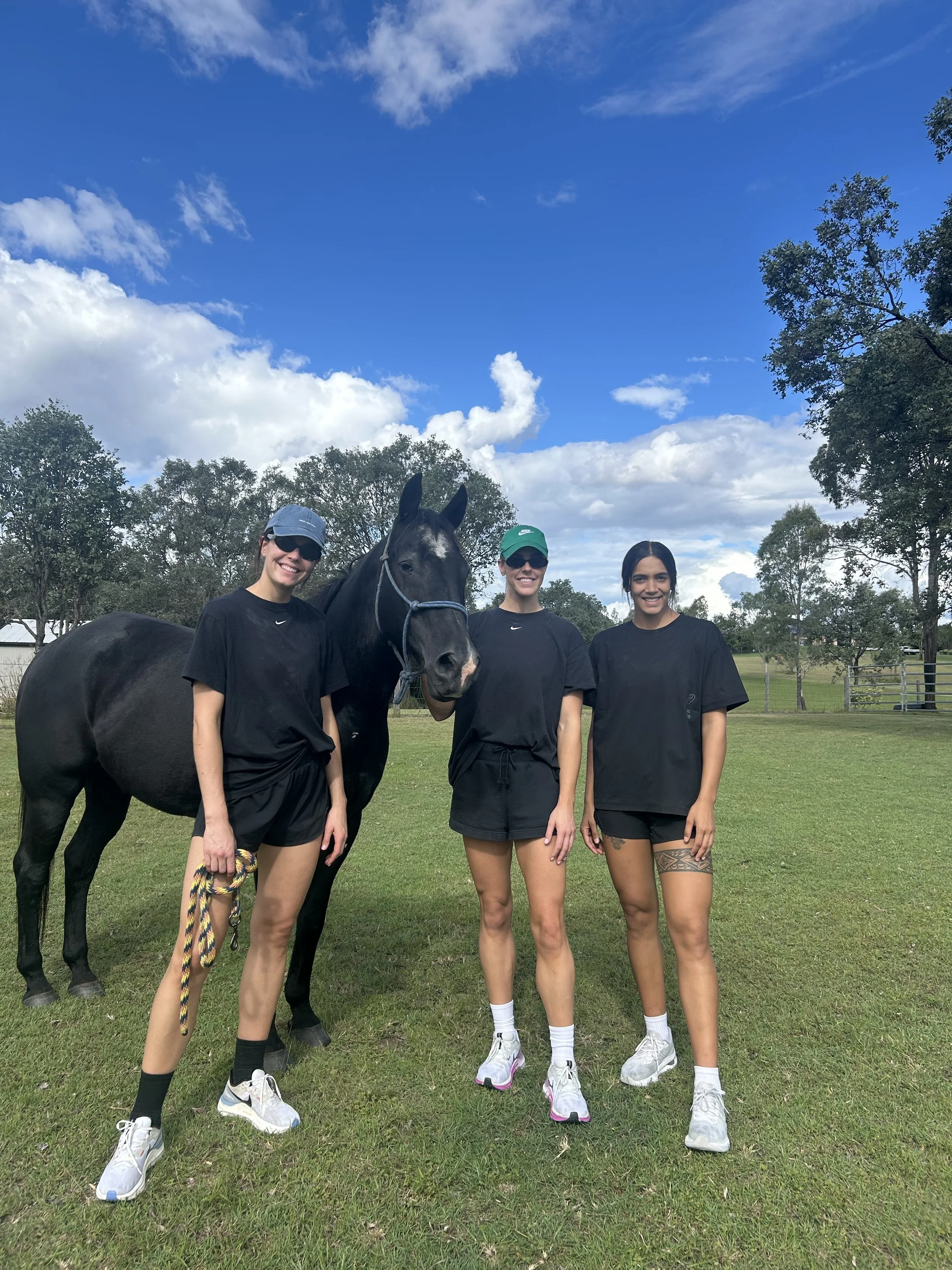 Three women standing outdoors on a grassy field with a horse. They are wearing black shirts and athletic shoes, and are smiling. The woman on the left is holding a multicolored leash attached to the horse. The sky is partly cloudy with blue sky visible. Trees and a fence are in the background.