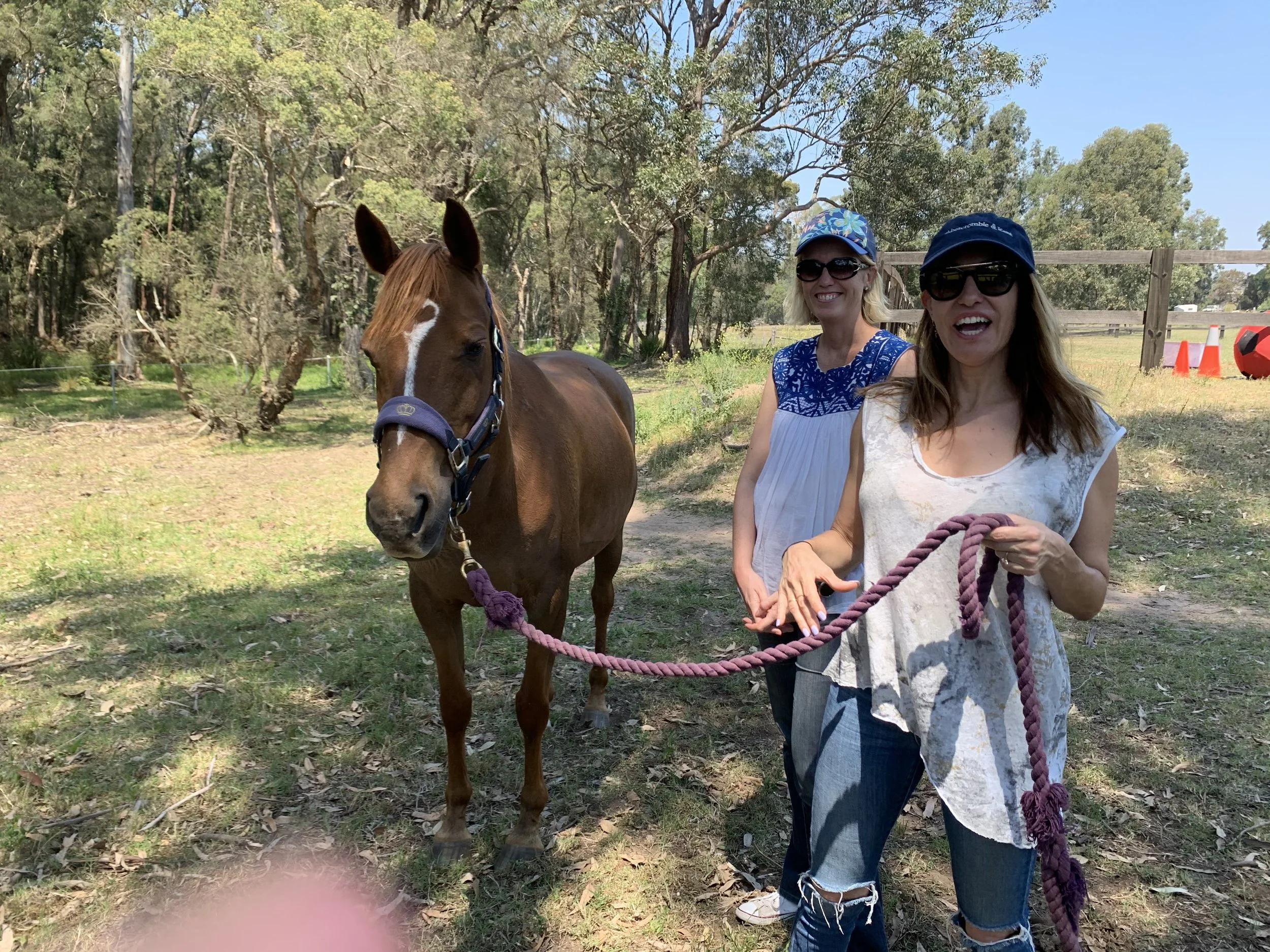 Two women standing outdoors with a horse on a leash, smiling and wearing sunglasses and hats, in a grassy area with trees and a wooden fence in the background.