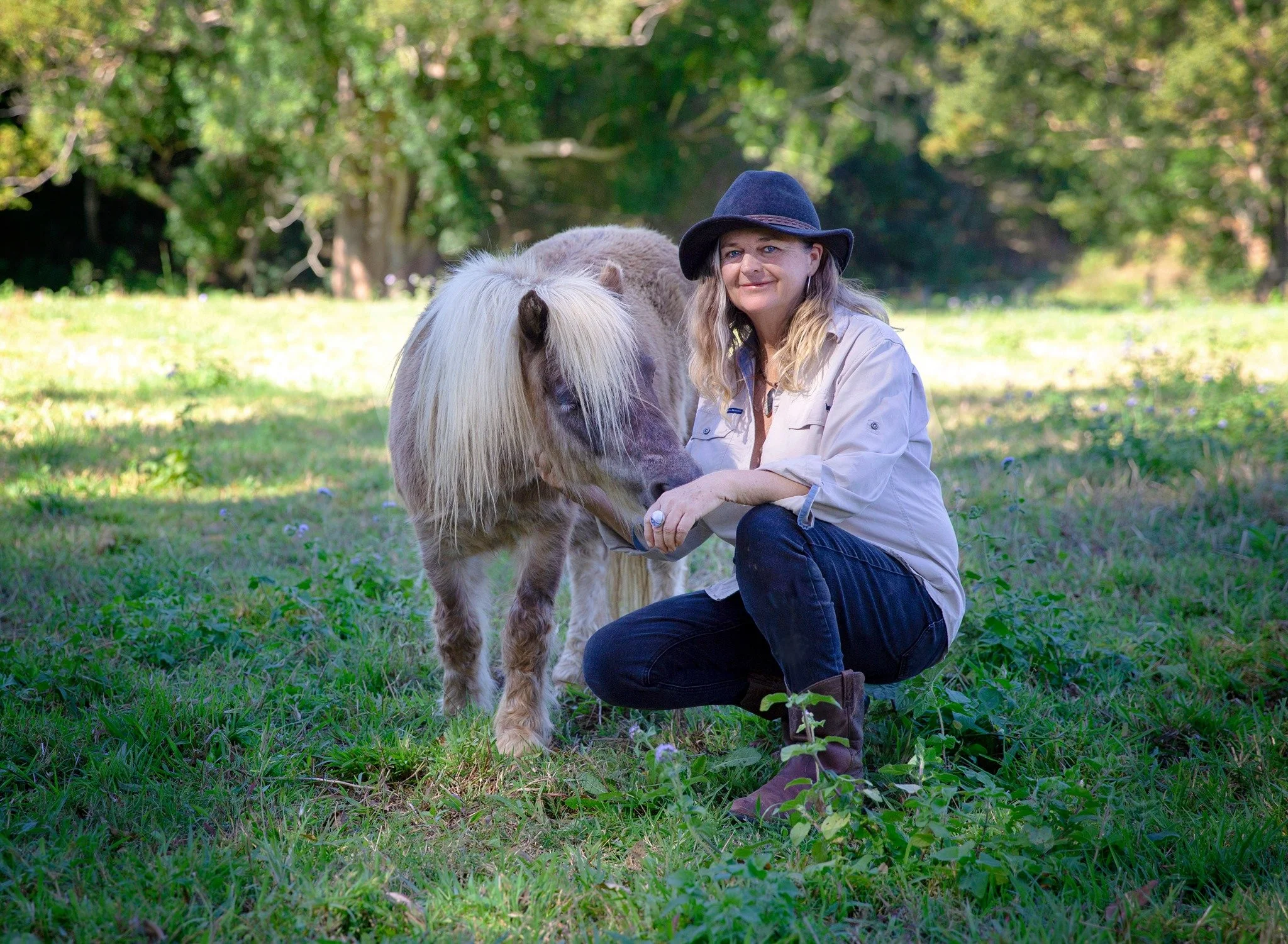 A woman kneeling on grass in a park during daytime, holding the head of a small horse or pony, with trees in the background.