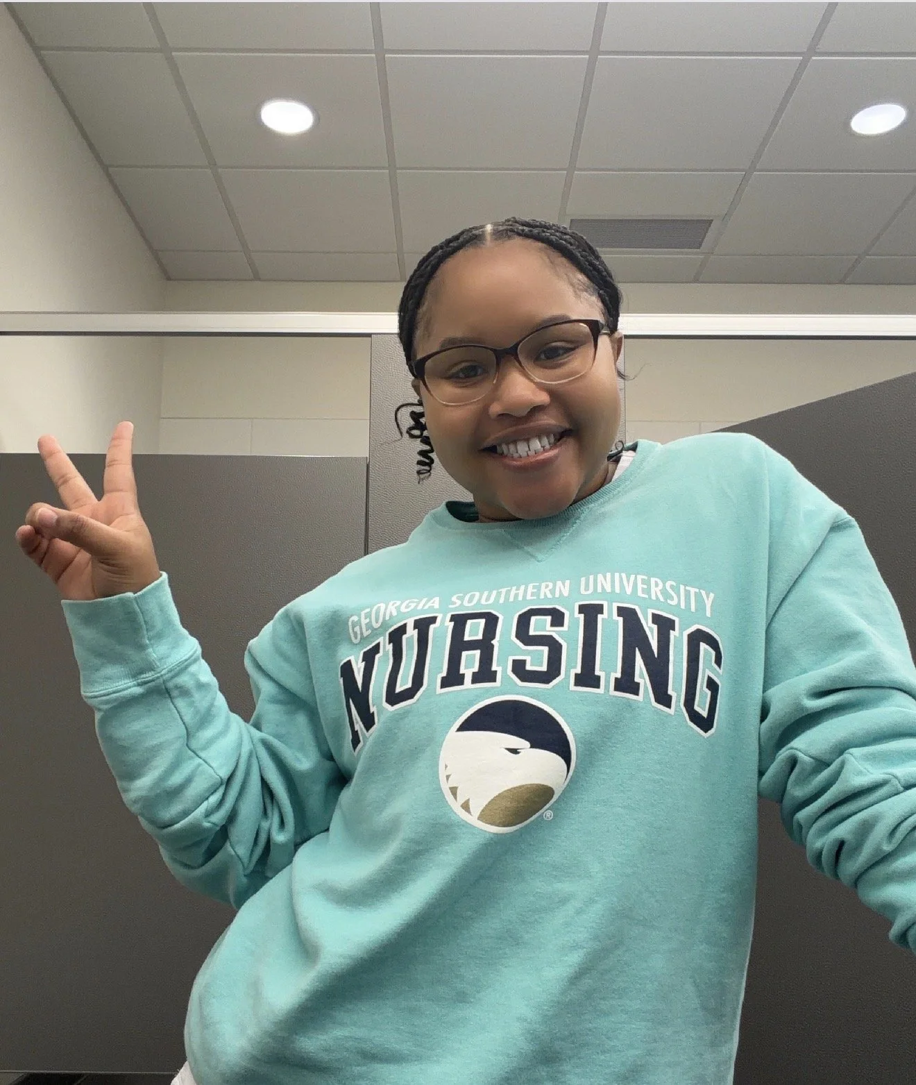 A young woman with glasses and braided hair smiling and making a peace sign, wearing a turquoise sweatshirt with Georgia Southern University NURSING logo.