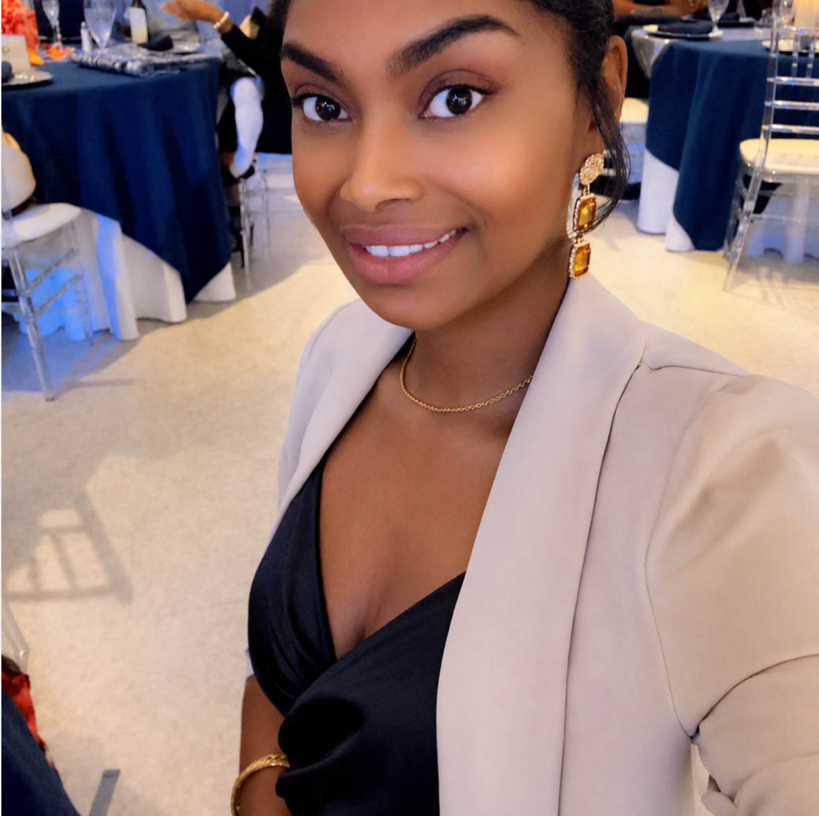 A woman taking a selfie at a formal event, dressed in a black dress, beige blazer, and gold jewelry, with decorated tables in the background.