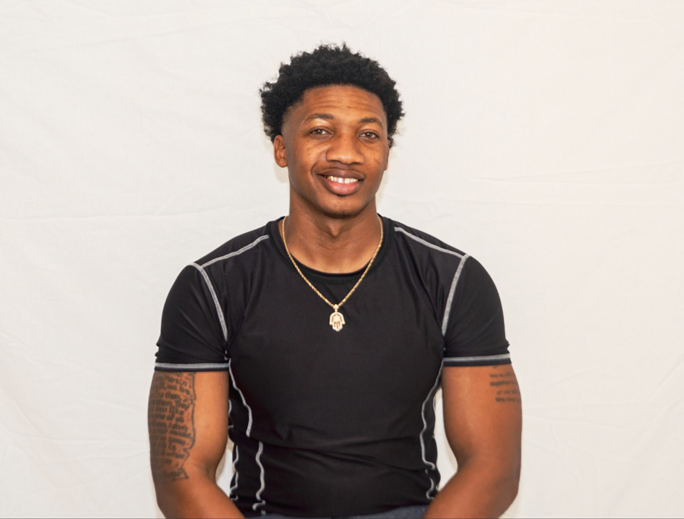 A young man with curly hair wearing a black athletic shirt with white trim and a gold chain necklace, smiling in front of a plain white background.