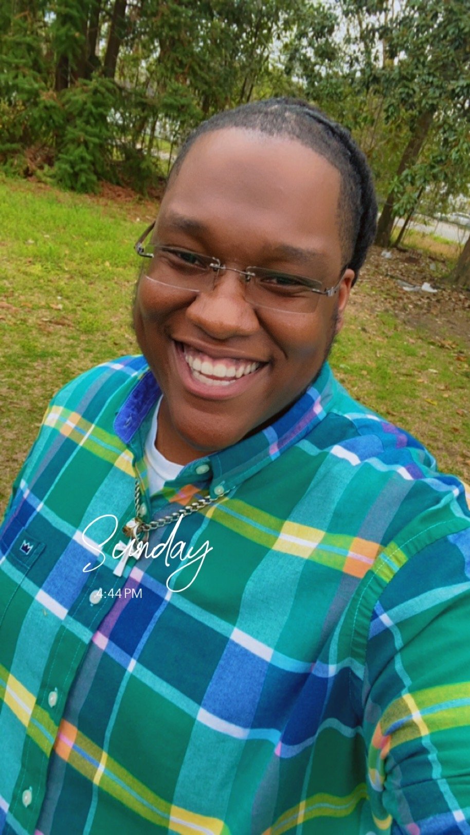 A smiling man wearing glasses and a colorful plaid shirt takes a selfie outdoors in a grassy area with trees in the background. The image shows the day of the week and time as Sunday at 4:44 PM.