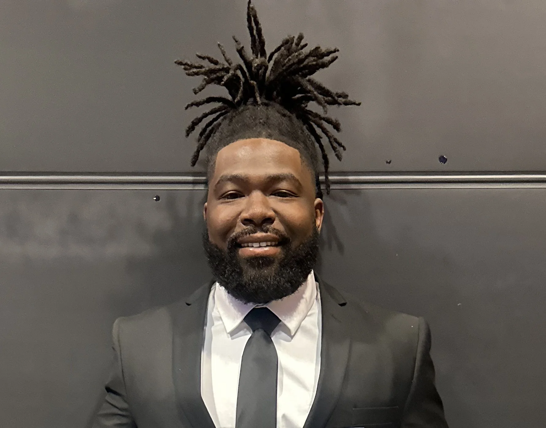 A man with dreadlocks styled upwards, wearing a dark suit, white shirt, and black tie, is smiling and standing against a dark background.