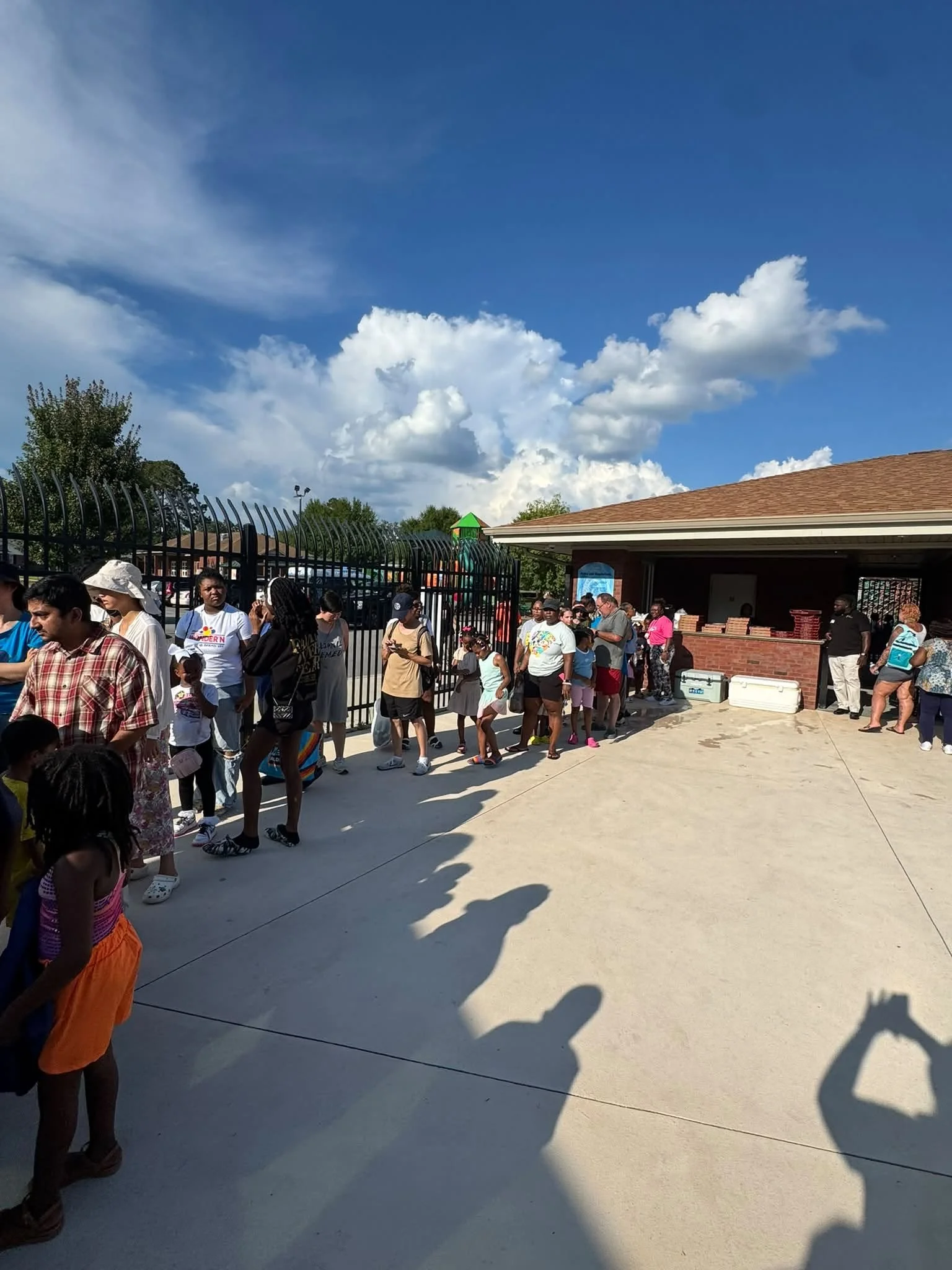 A diverse group of people, including children and adults, standing in line outside near a brick building with a parking lot entrance gate under a partly cloudy blue sky.