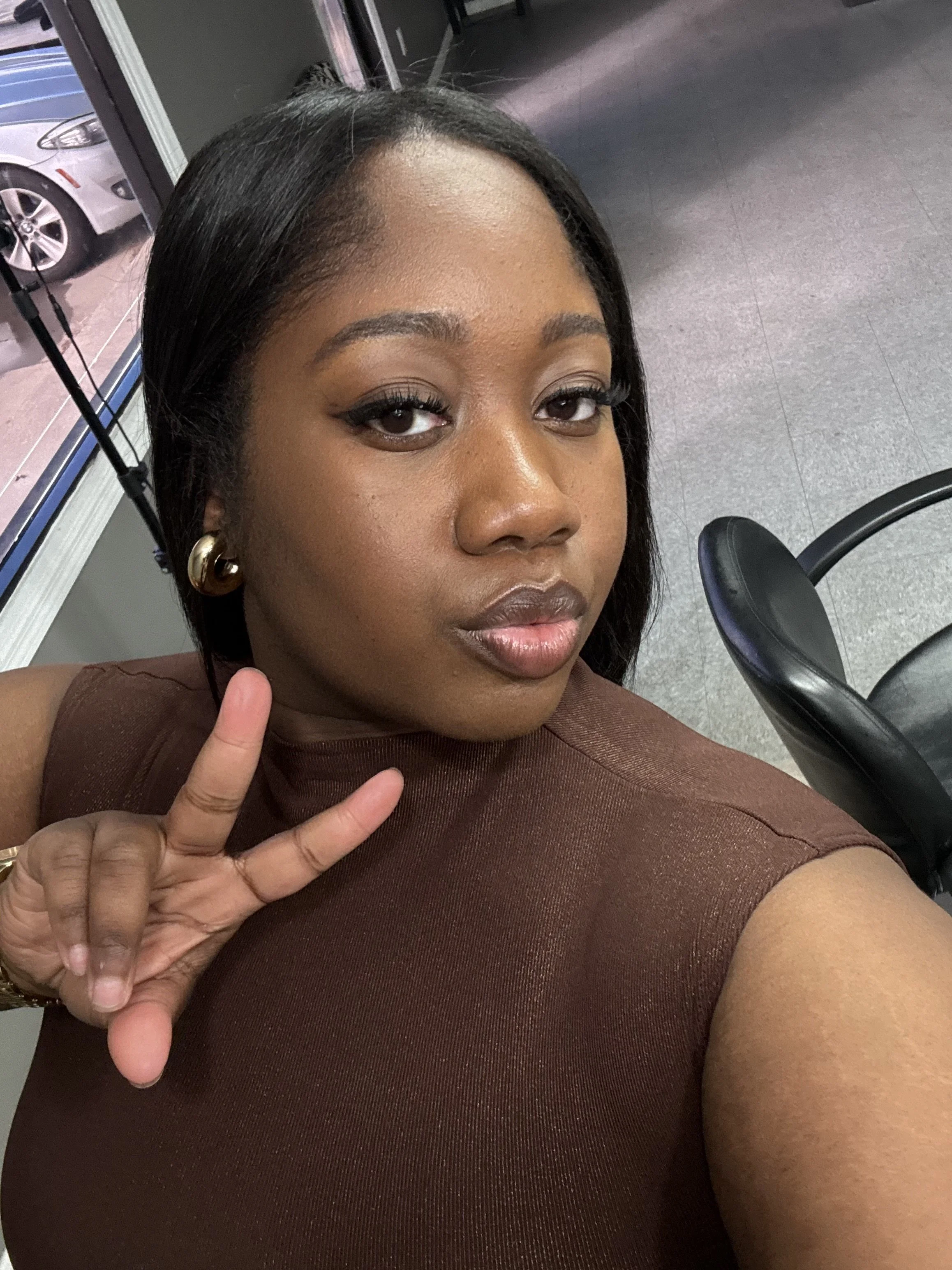 A woman with dark hair and makeup posing for a selfie at what appears to be an airport or train station, making a peace sign with her fingers and wearing a brown sleeveless top.
