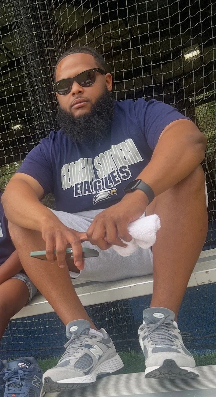 A man sitting on a bench wearing a Georgia Southern Eagles t-shirt, sunglasses, and sneakers, holding a smartphone and a white towel, with a background of a sports fence and a person partially visible.