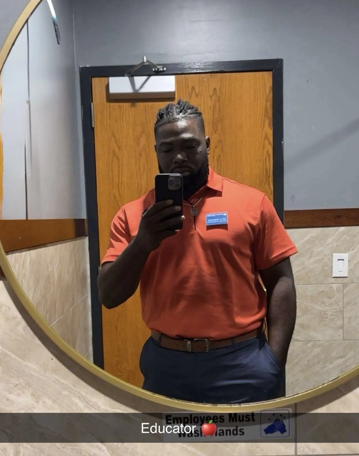 A man taking a mirror selfie in a bathroom, wearing an orange polo shirt and dark gray pants, with a wooden door and beige tiled walls in the background. There is a sign on the mirror that says "Employees Must Wash Hands" and has a red apple icon and a blue handwashing icon.