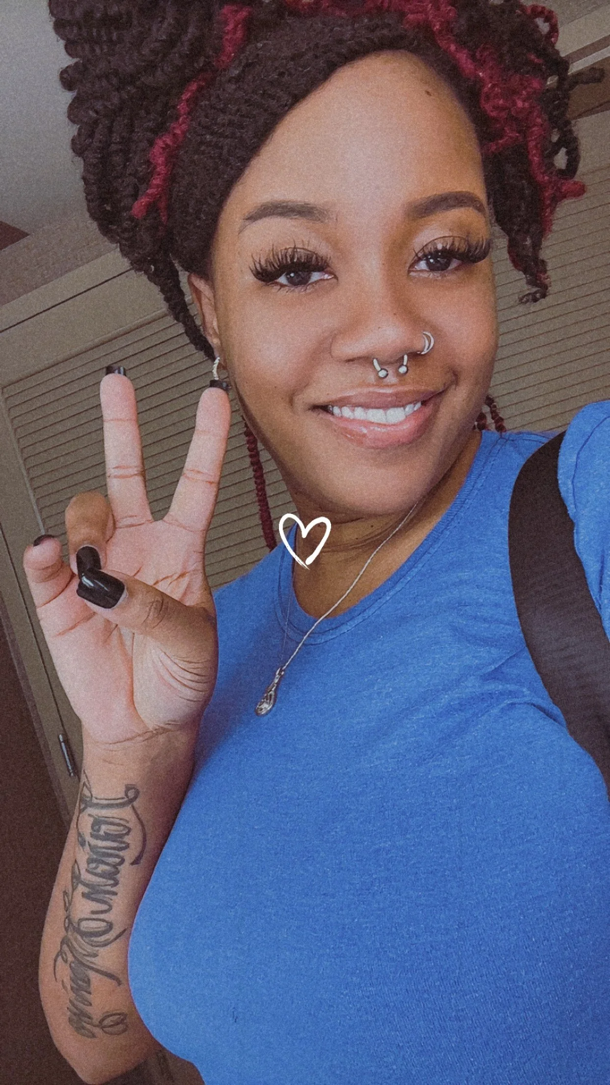 A woman with red-brown curly hair styled in twists, making a peace sign with her hand, wearing a blue shirt, jewelry, and tattoos on her arm, smiling at the camera.