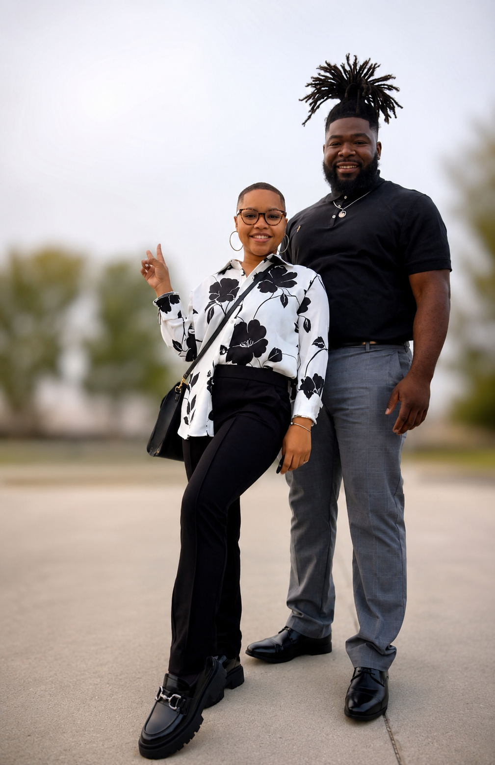 A smiling man and woman standing outdoors on a paved area, with the man showing dreadlocks and wearing a black shirt and gray pants, the woman wearing glasses, a white blouse with black floral pattern, and black pants.