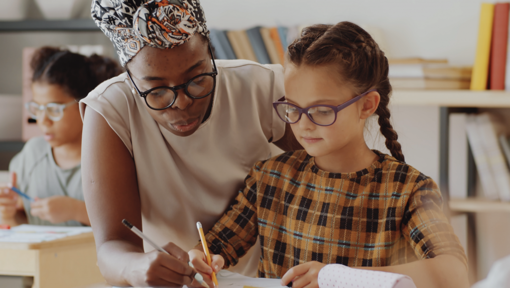 A female teacher helping a young girl with glasses in a classroom, with other students in the background.
