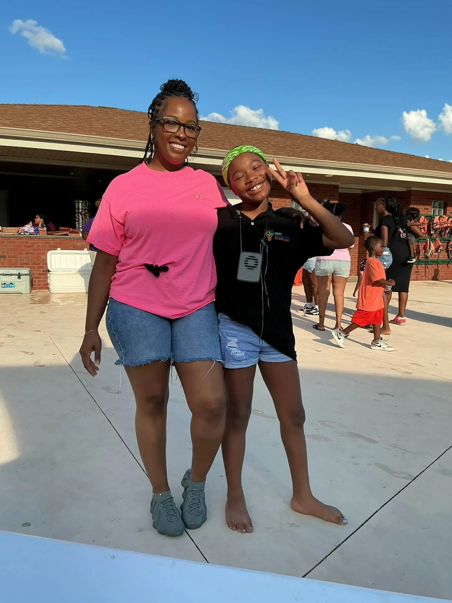 A woman and a young girl posing together for a photo outdoors on a sunny day. The woman is wearing a pink T-shirt, denim shorts, and sneakers. The girl is wearing a black shirt with a colorful logo, blue shorts, flip-flops, and a green headscarf, making a peace sign with her fingers.
