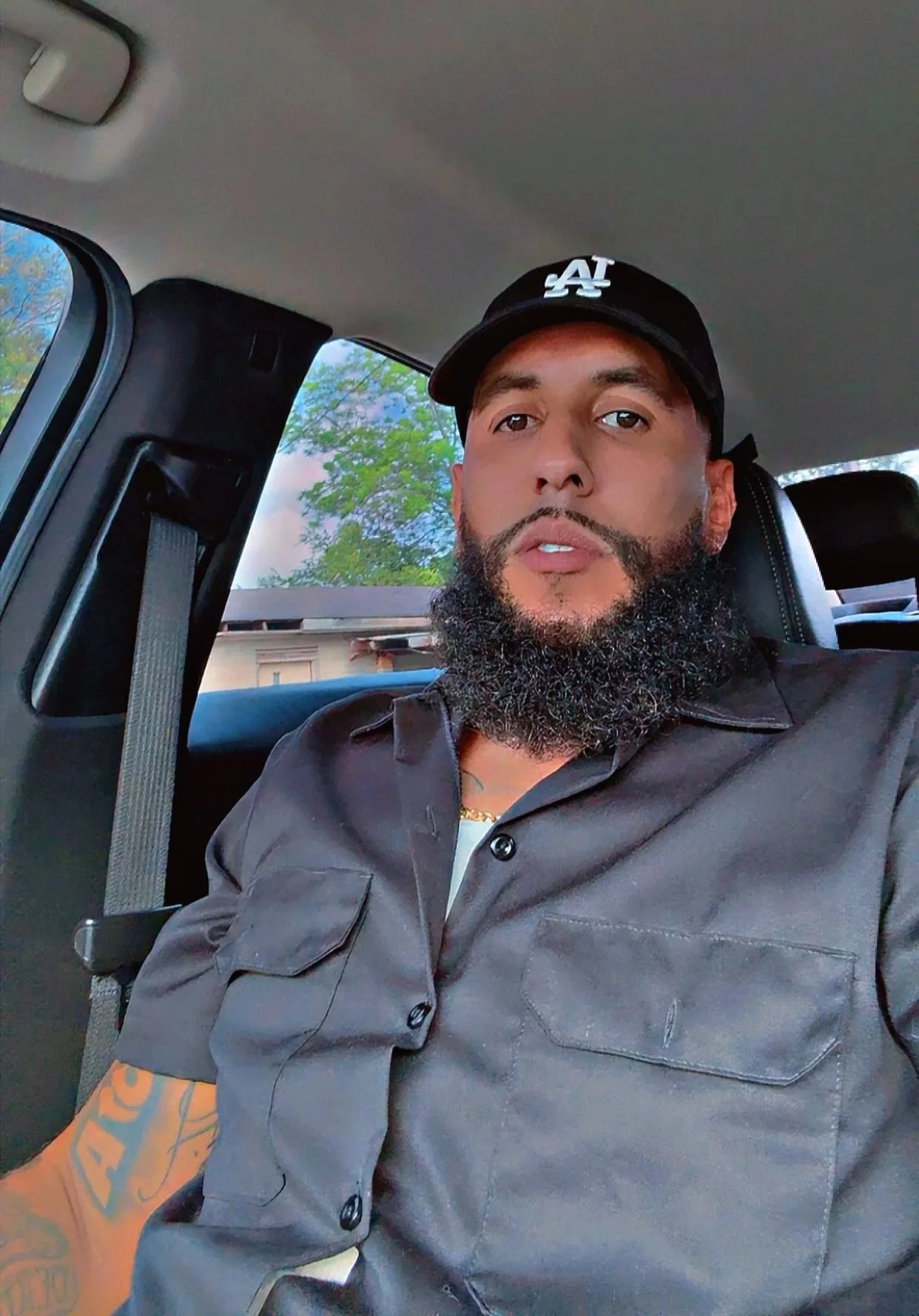 A man with a beard wearing a black LA baseball cap and a dark button-up shirt sitting in a vehicle, with trees and sunlight visible through the window behind him.
