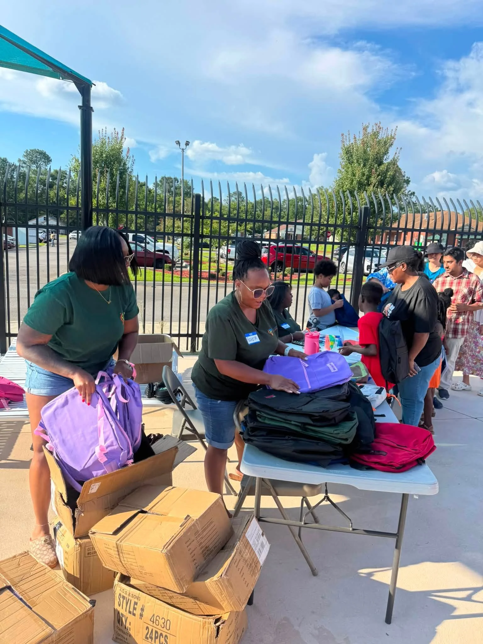 People gathering outdoors behind a black metal fence, with tables filled with backpacks, boxes, and supplies, under a blue sky with scattered clouds.