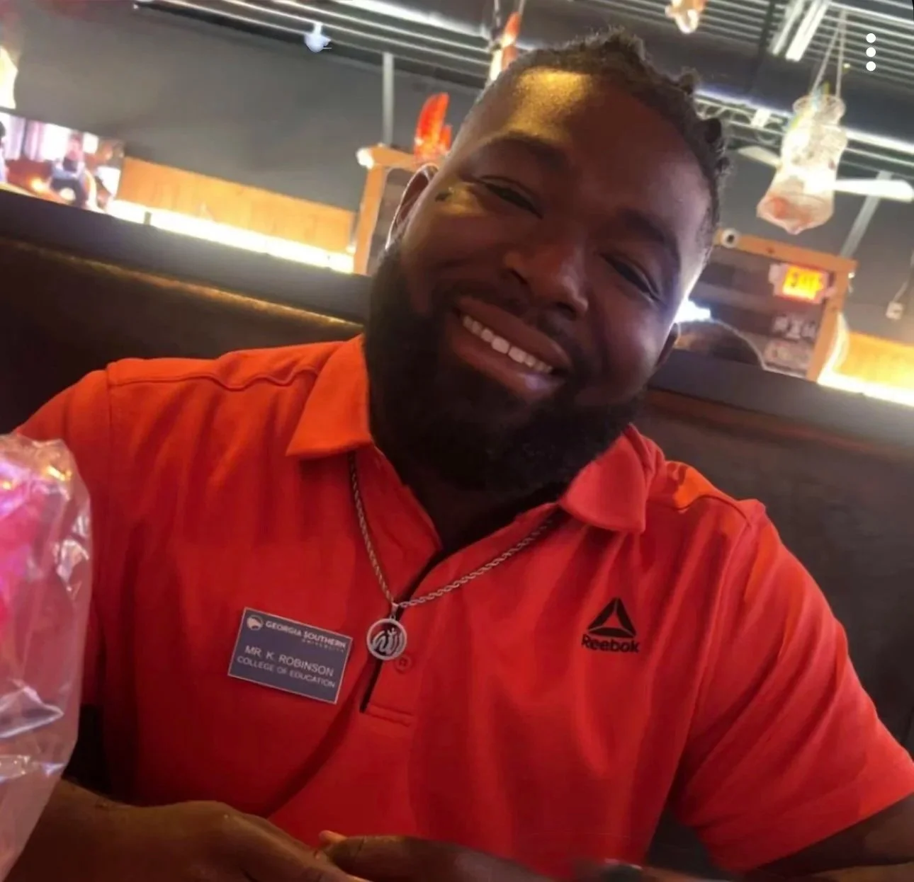 A smiling man with a beard, wearing an orange Reebok shirt and a name badge, sitting in a restaurant or cafe.